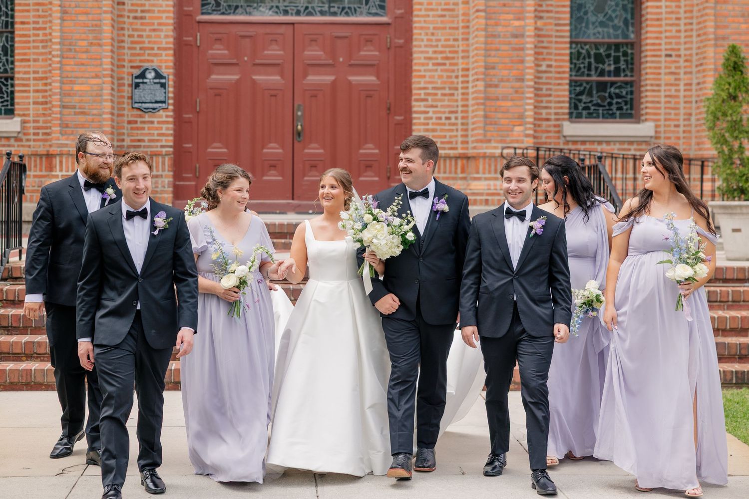 Wedding party walks together outside brick church with bride in white gown and bridesmaids in lavender.