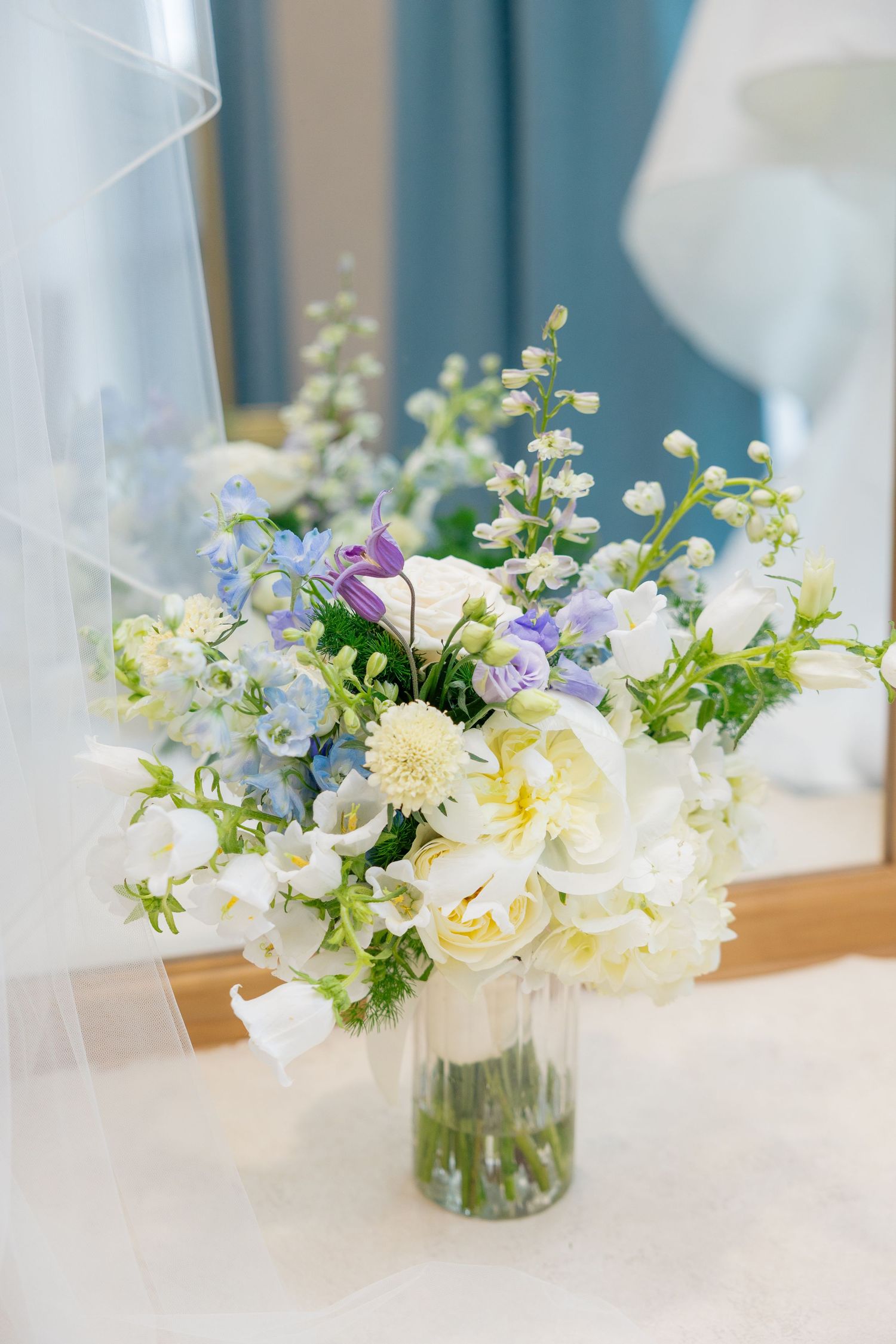 Bridal bouquet of white, blue, and purple flowers in glass vase on dressing table before wedding.