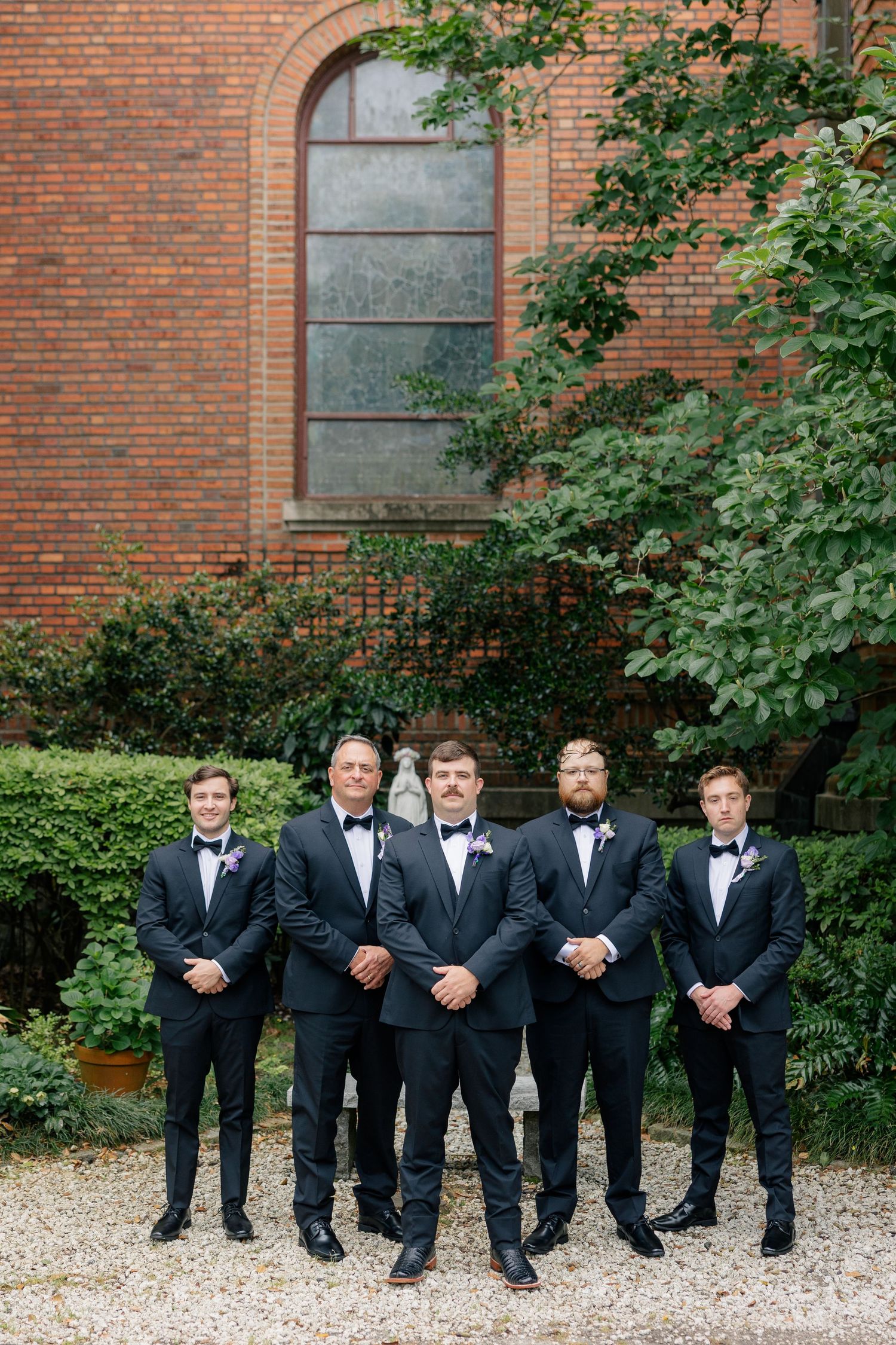 Five groomsmen in matching black tuxedos pose together outside a brick building with lush greenery.