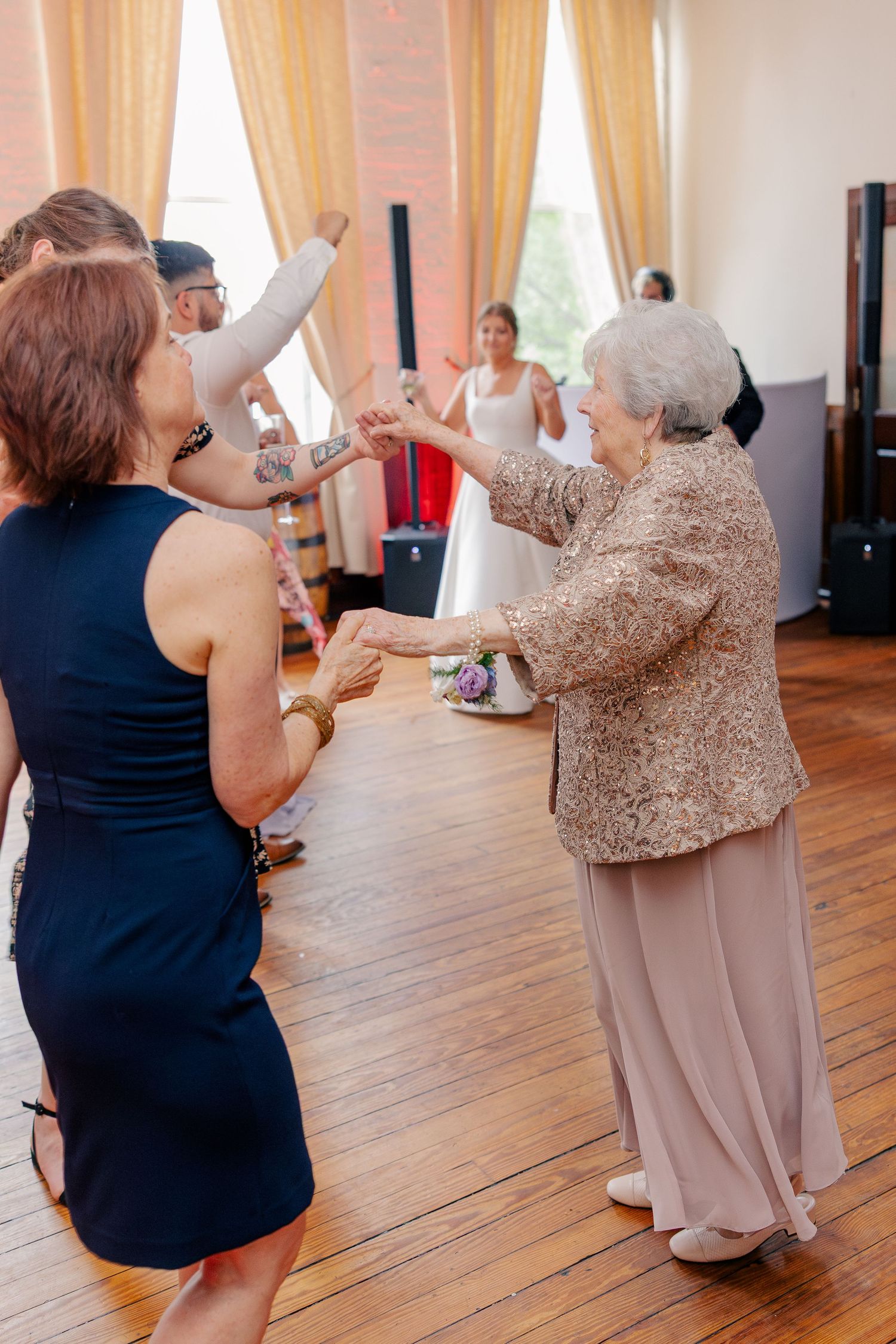 Elderly woman and guests dancing joyfully on wooden dance floor at wedding reception.