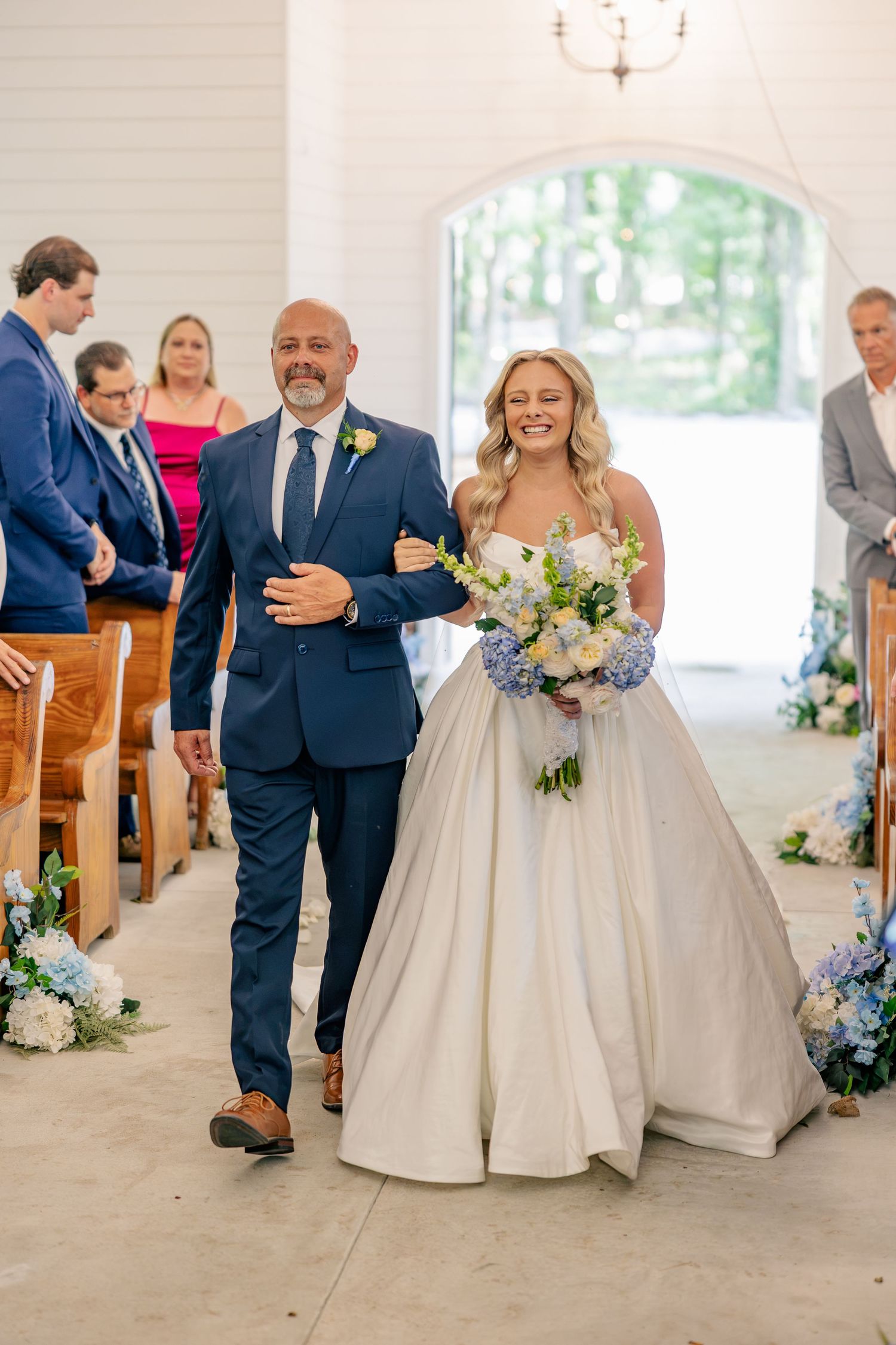 Smiling bride walks down chapel aisle with bouquet of blue and white flowers escorted by father.