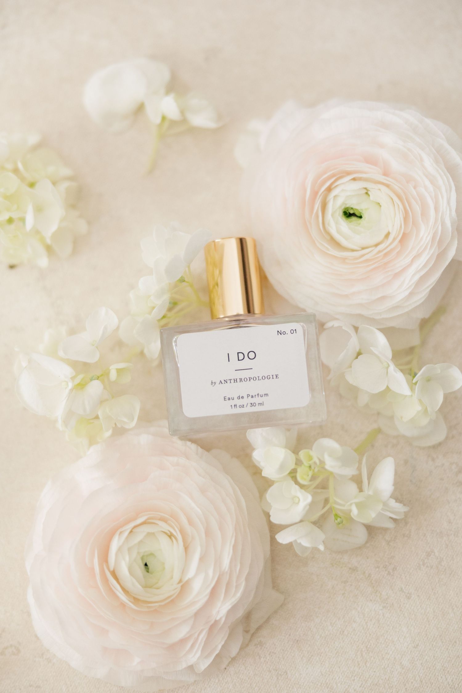Anthropologie I Do eau de parfum bottle surrounded by soft pink ranunculus and white hydrangea blooms.