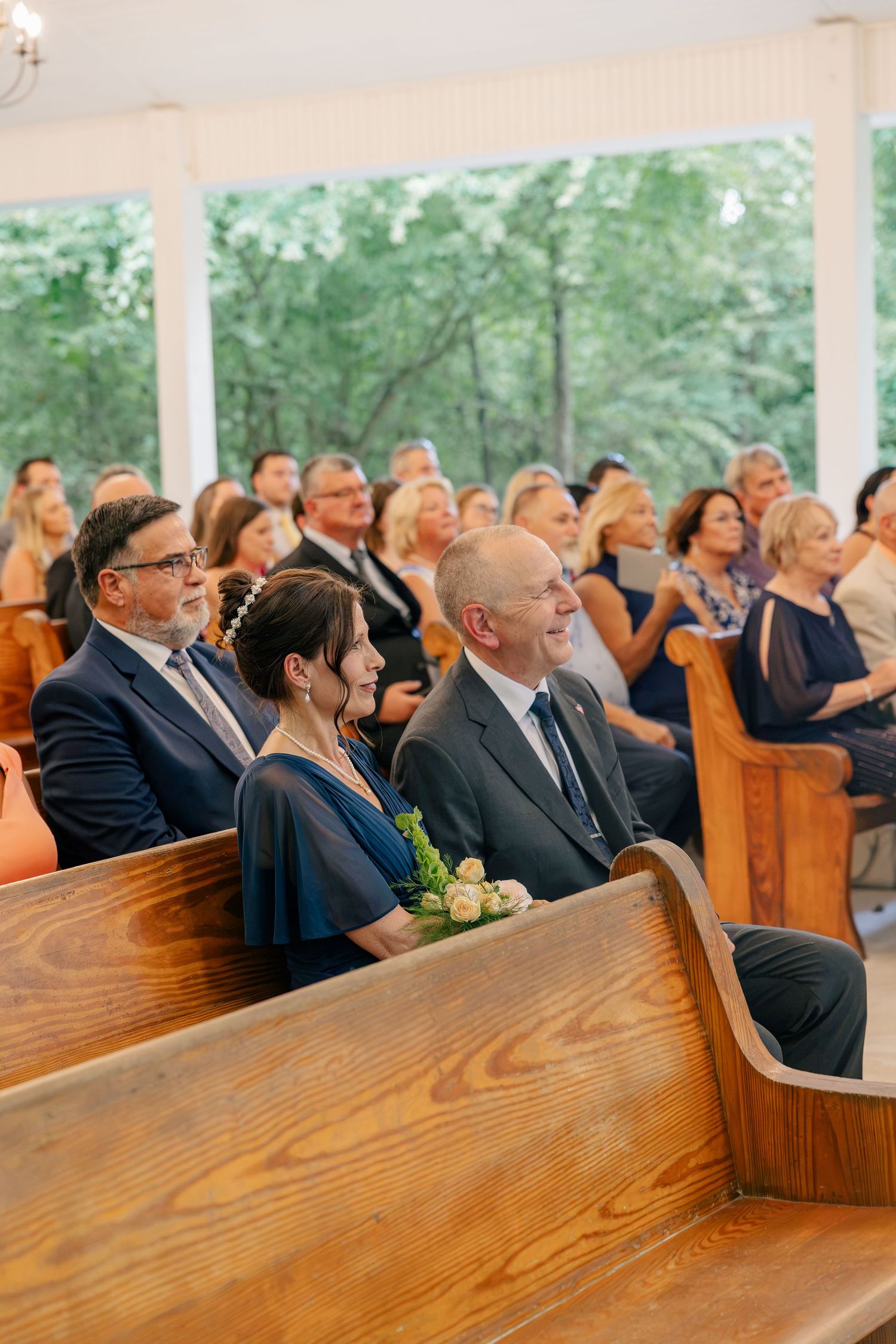 Guests seated in wooden church pews smile during an outdoor wedding ceremony, with lush green trees visible through large windows.
