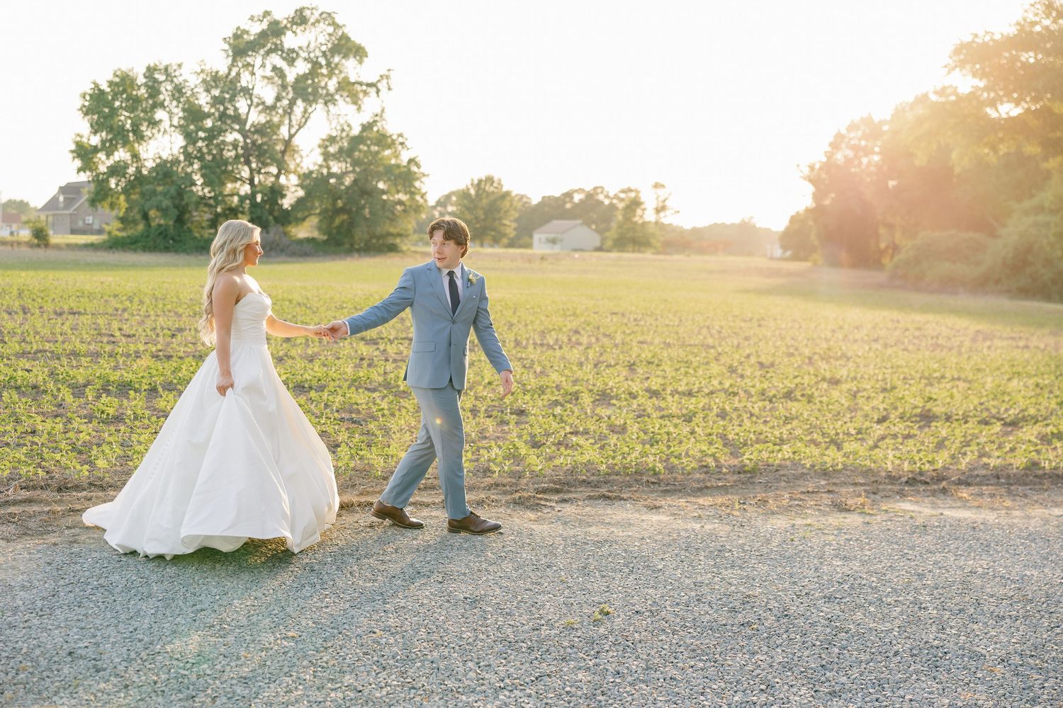 Bride and groom hold hands walking through sunlit field during golden hour wedding portraits.