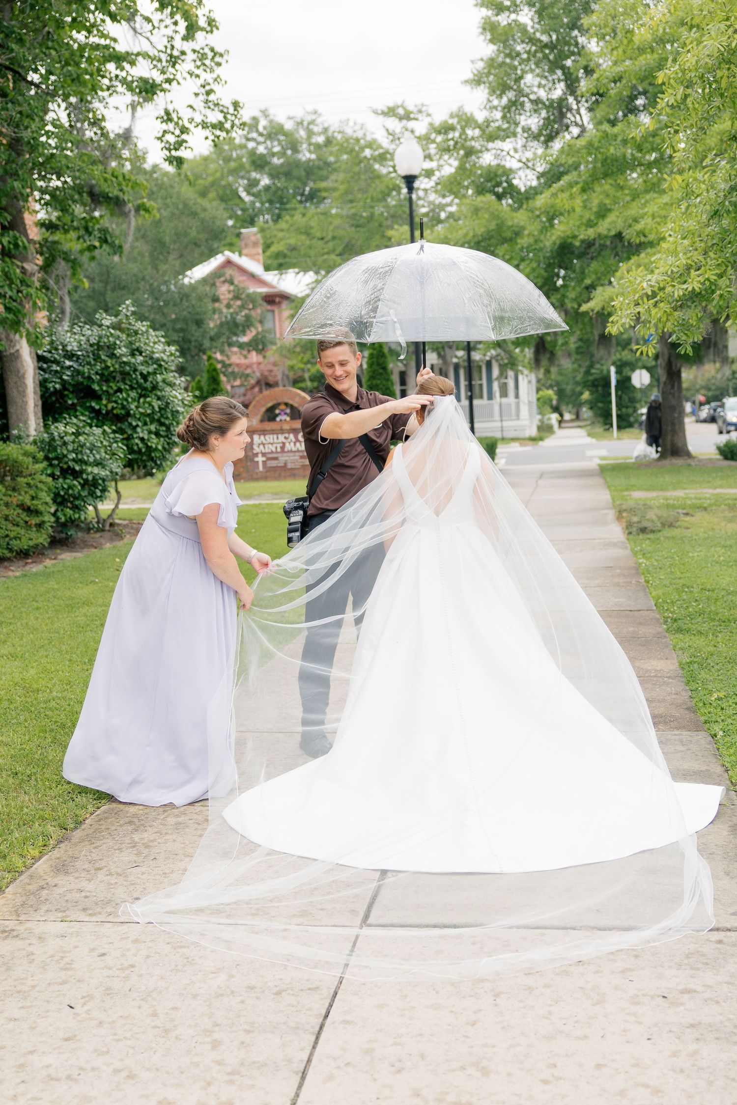 A bride in a white gown and veil being assisted by two people on a sidewalk, one holding a clear umbrella outdoors.