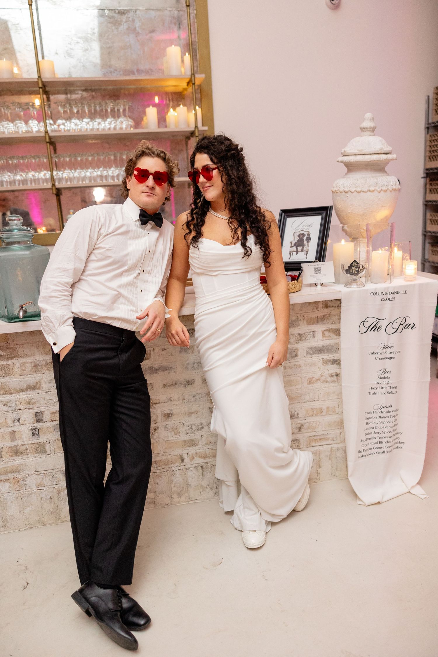 Wedding couple wearing fun red sunglasses pose casually against a brick wall with glassware display.