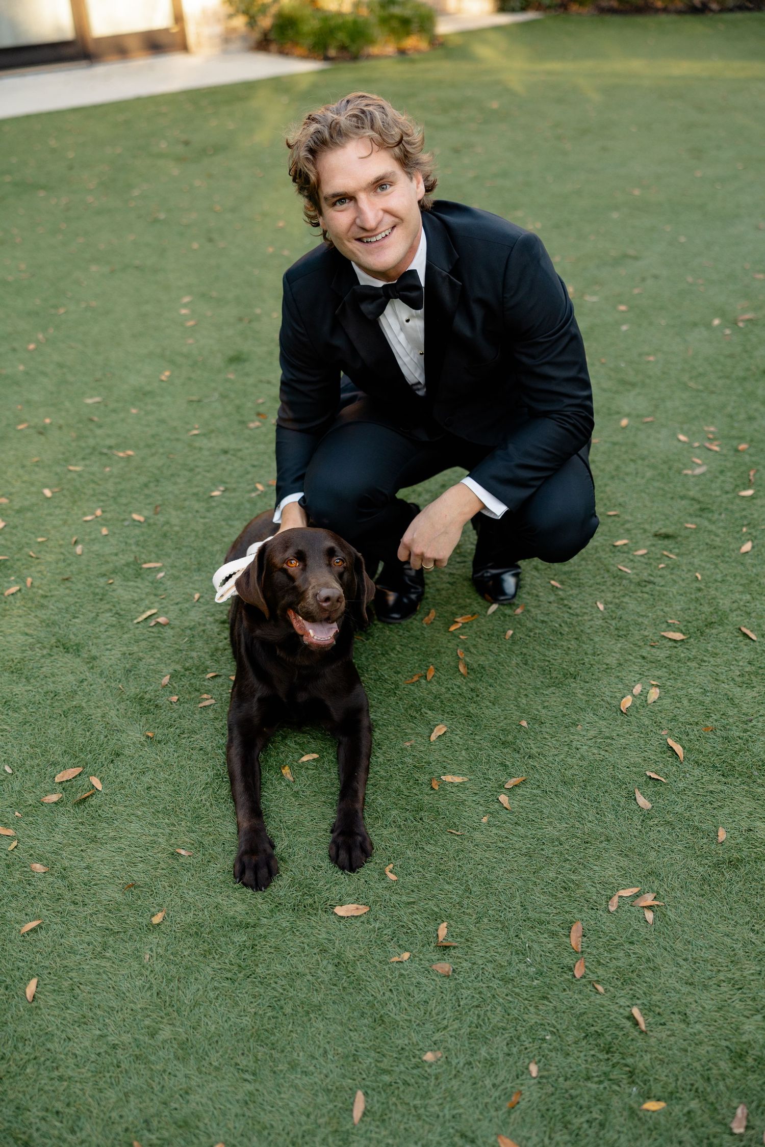 A person in a formal suit kneels beside a happy chocolate Labrador on green grass.