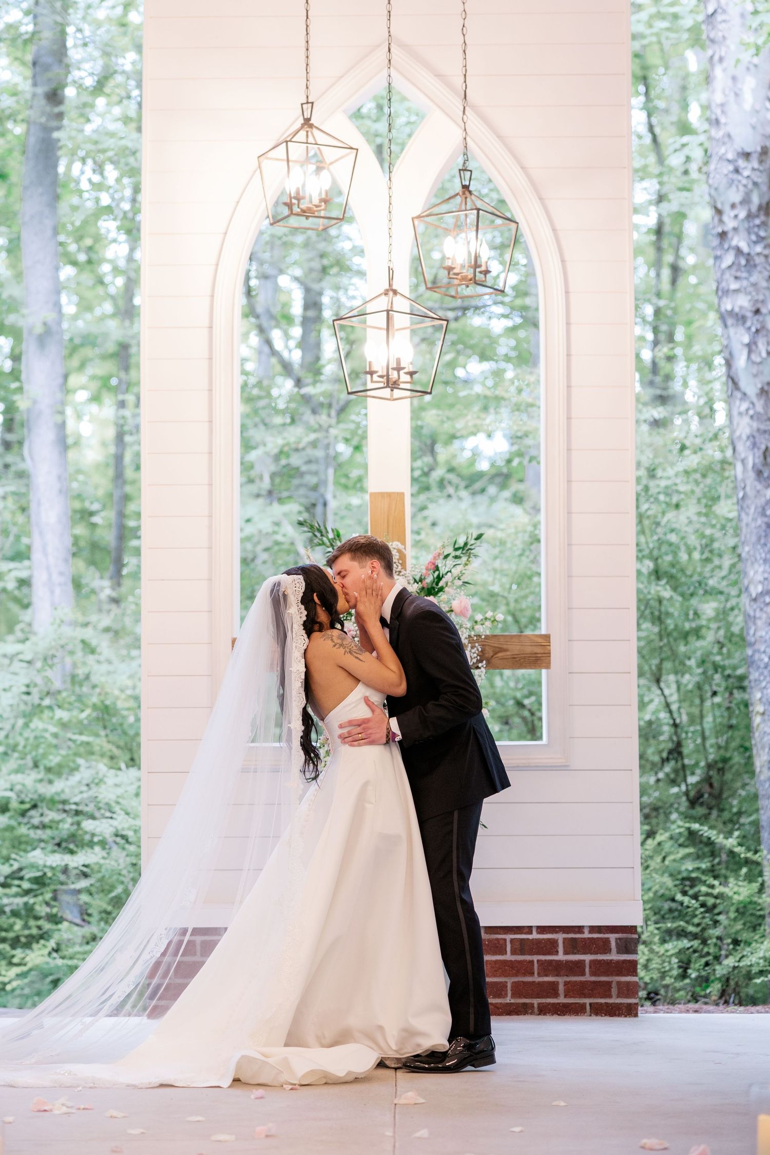 Romantic wedding couple sharing a kiss in a white columned outdoor venue with forest backdrop.