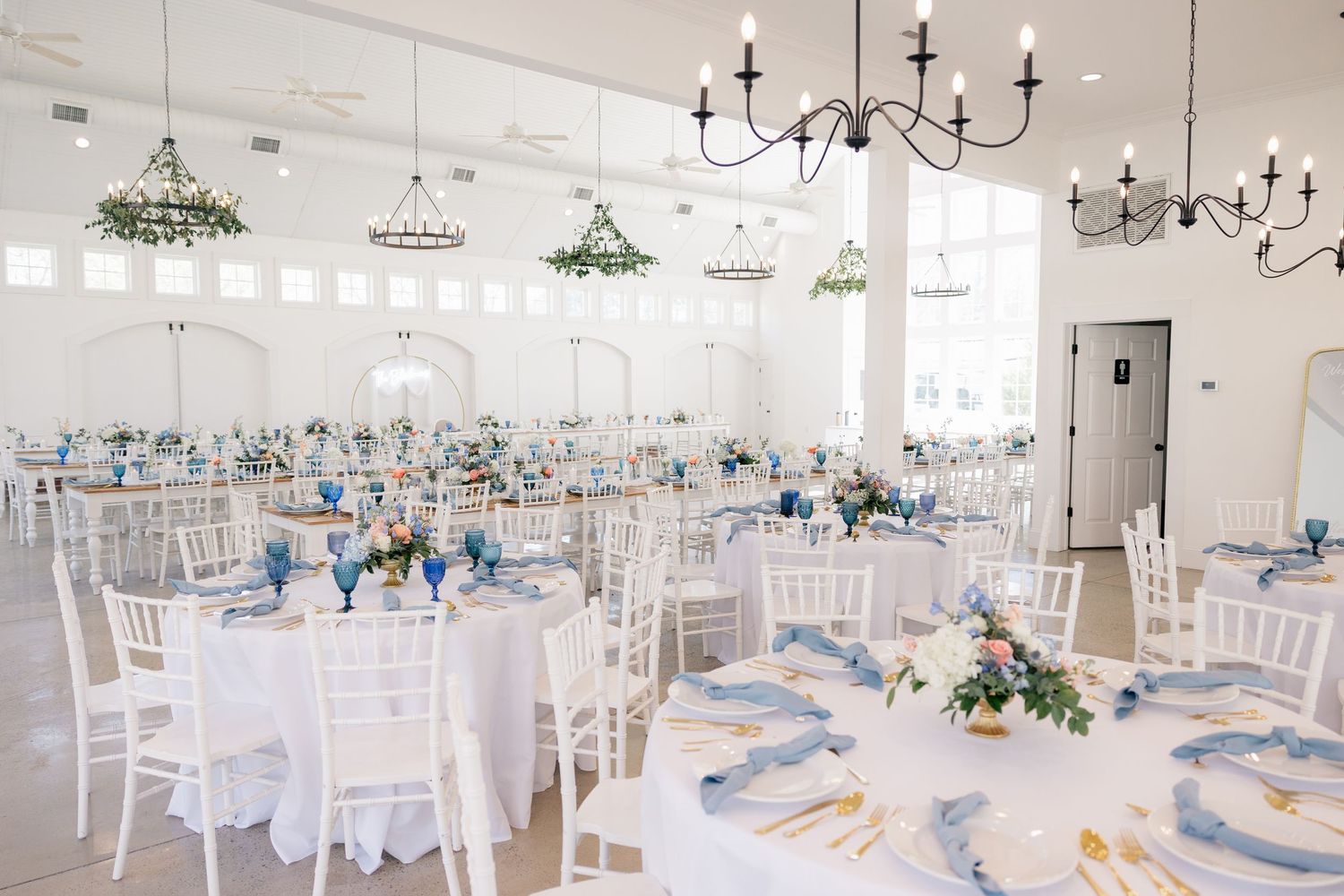 Elegant white reception hall with chandeliers and white chairs set for a formal wedding celebration.
