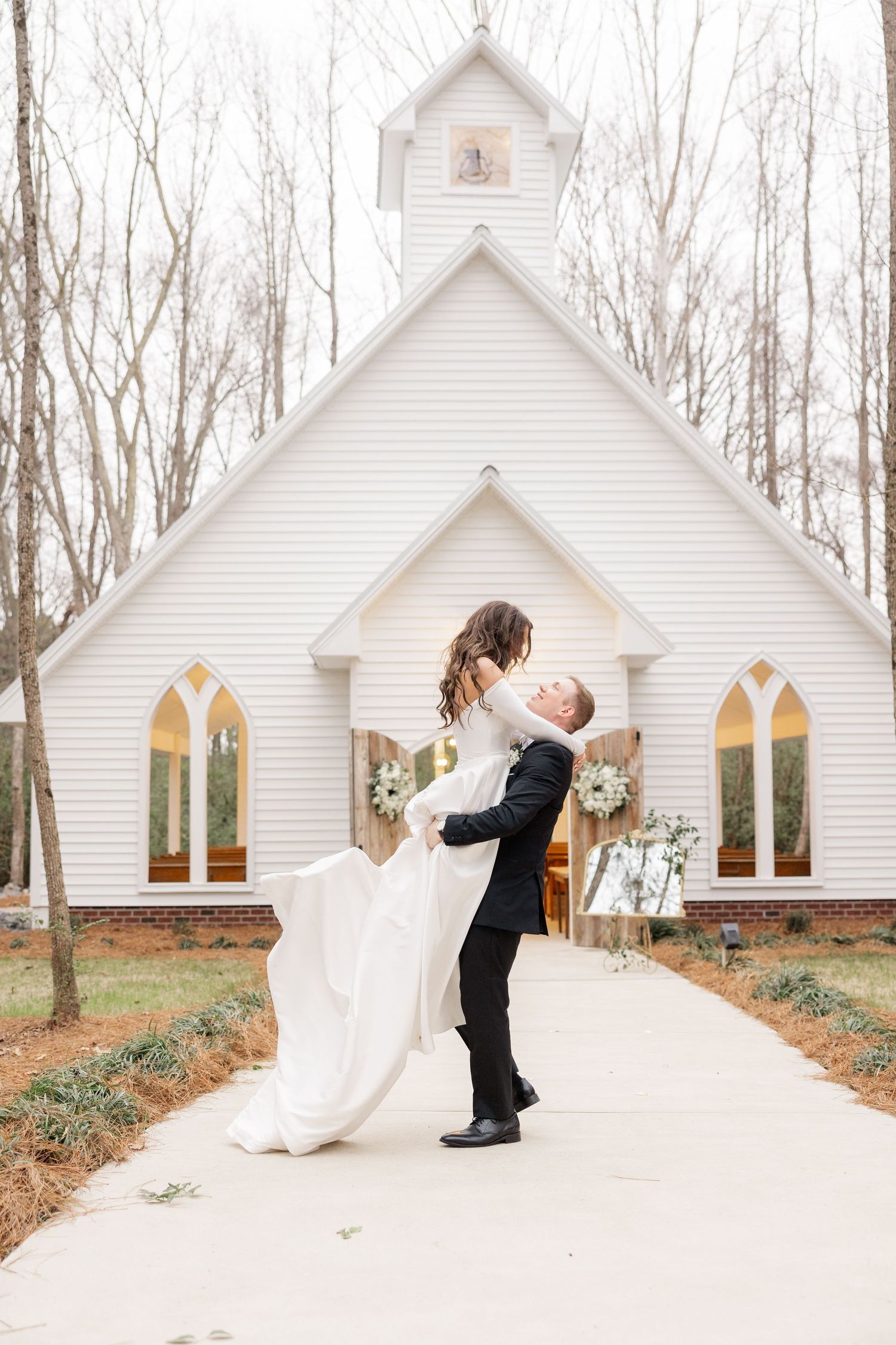 A romantic wedding photoshoot outside a classic white chapel surrounded by bare winter trees.