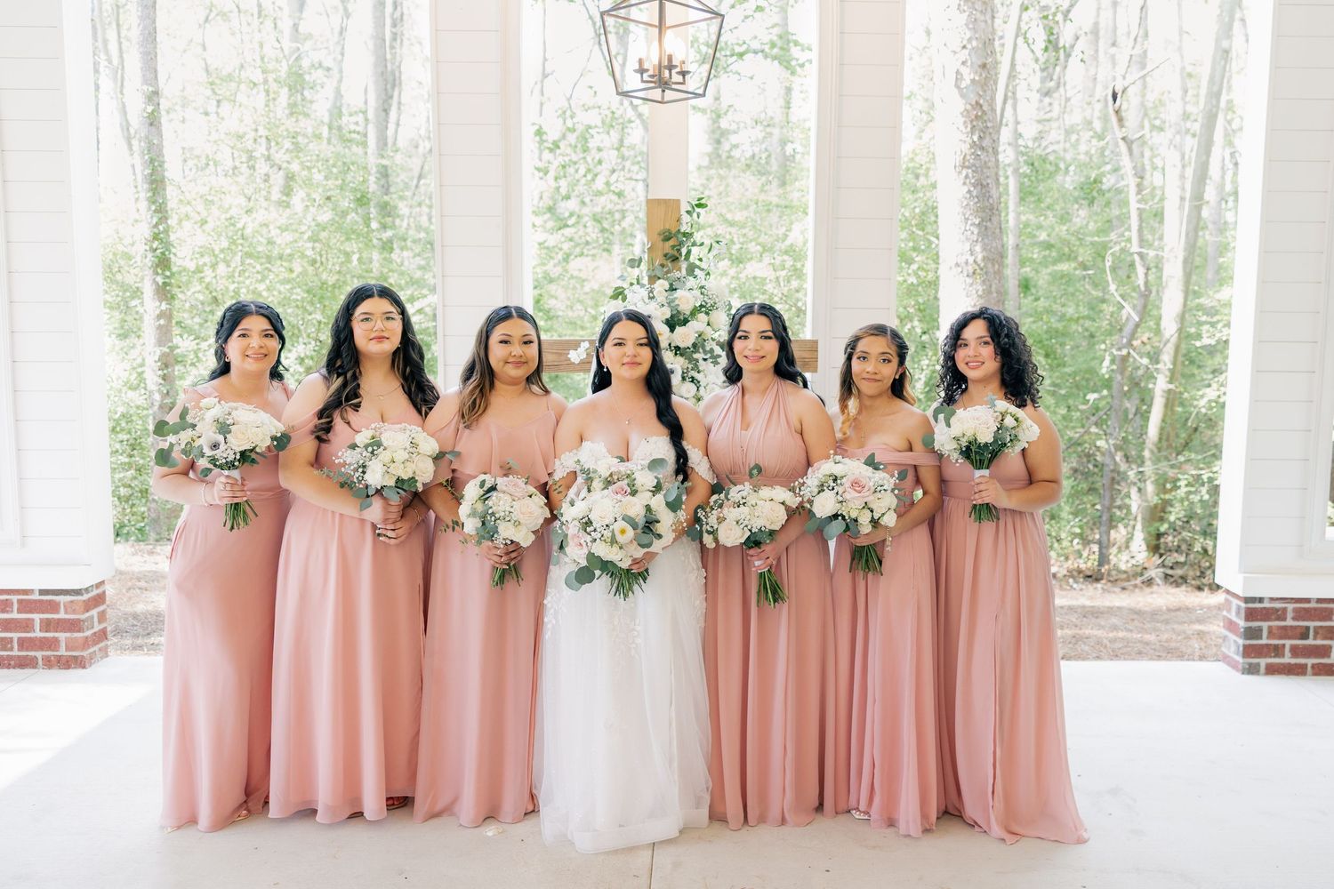 Bridesmaids in dusty rose dresses stand together holding white bouquets during an outdoor wedding ceremony.