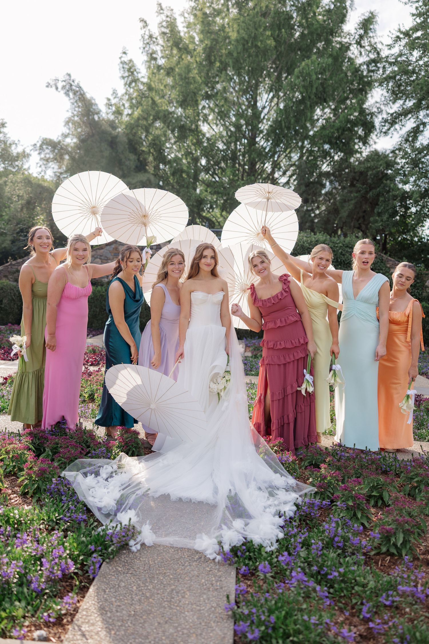 Wedding party posing with white parasols among purple flowers in a garden setting.