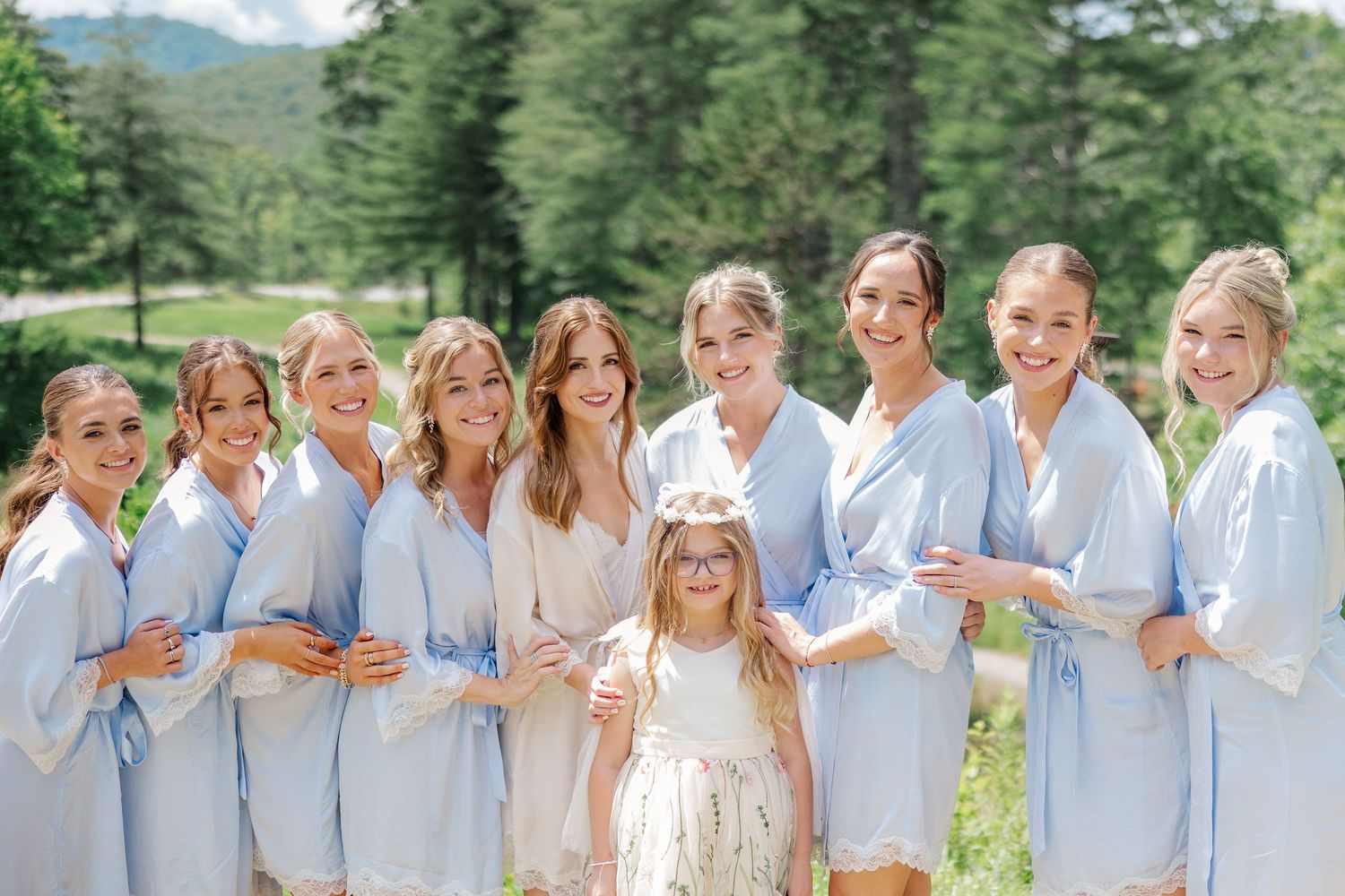A group of people wearing white and beige outfits pose together outdoors with mountains and trees in the background.