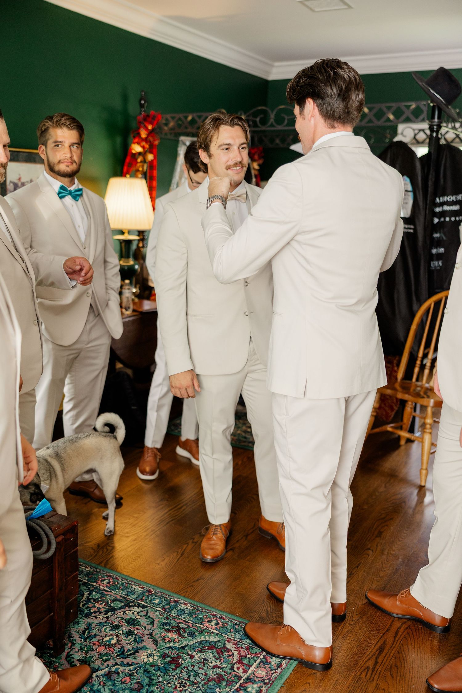 Groomsmen in light colored suits and blue bow ties gather in a room with green walls and hardwood floors.