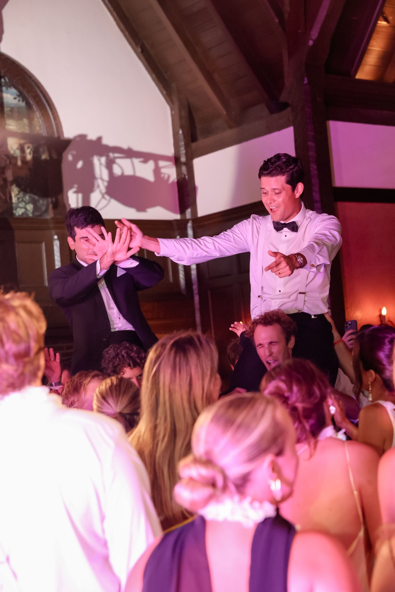 Two men dance energetically on elevated platform surrounded by wedding guests on lit dance floor.