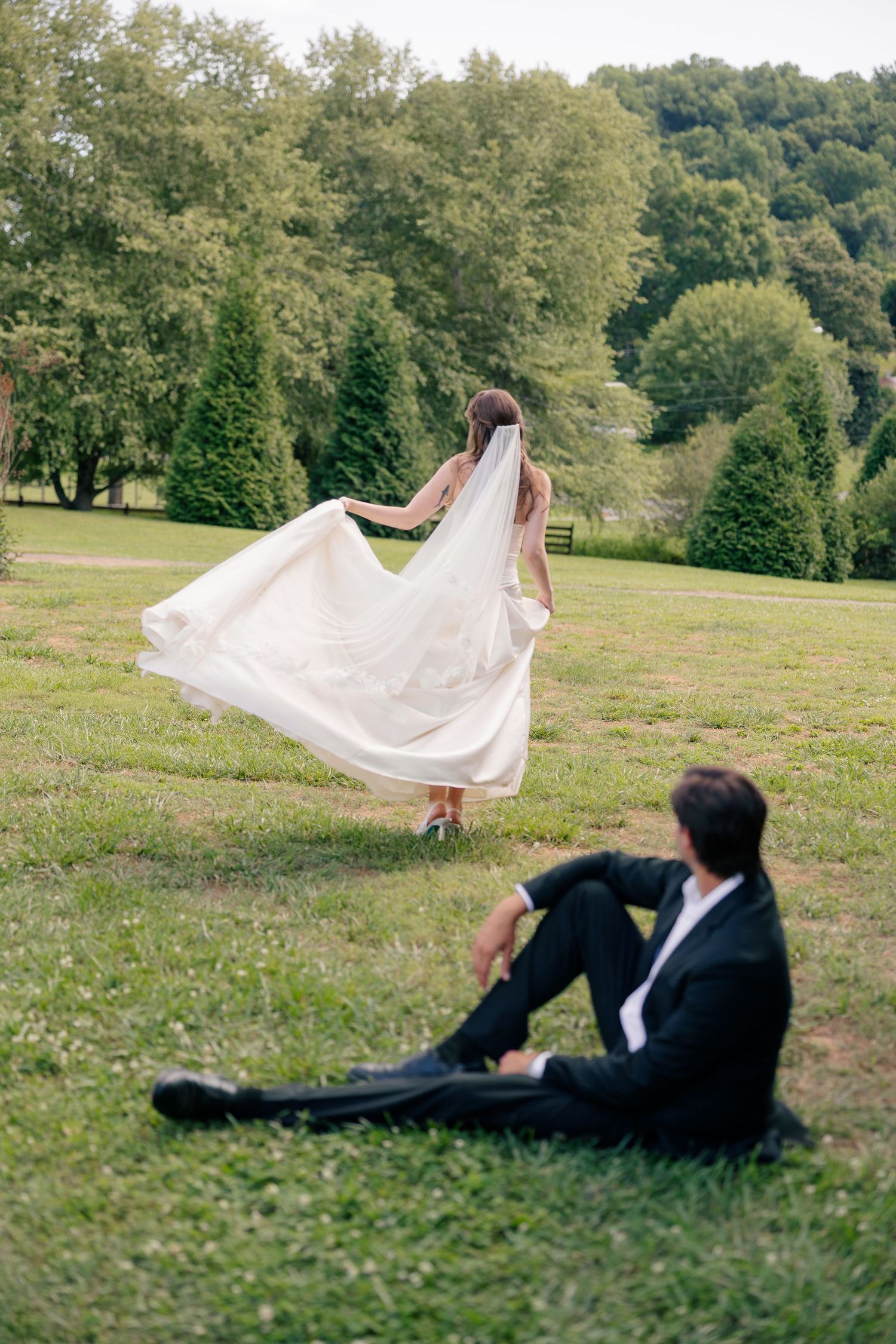 Bride in flowing gown and veil runs across green hillside as groom relaxes on grass.