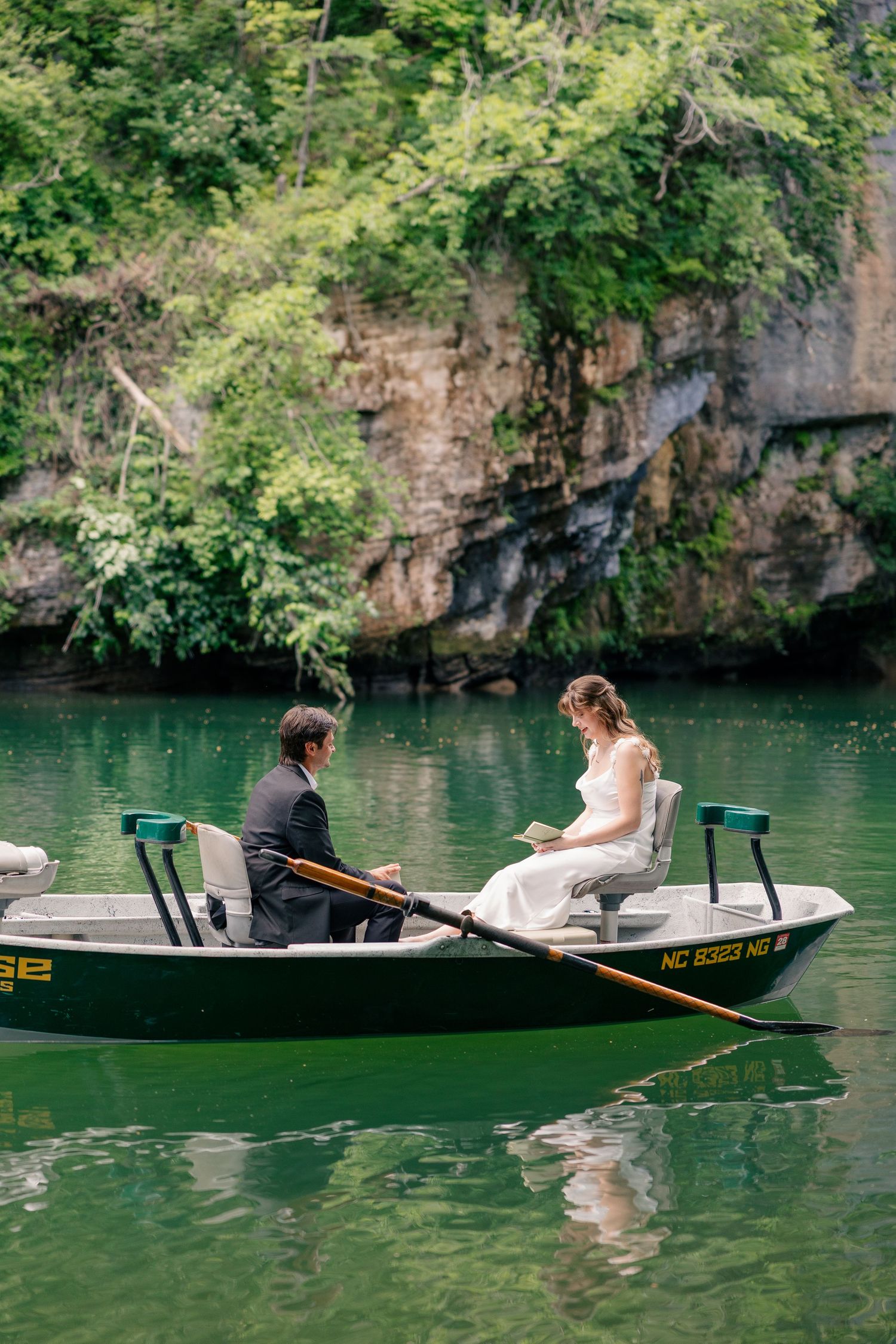 Bride and groom share intimate moment reading vows in a green rowboat on a serene lake surrounded by lush cliffs.