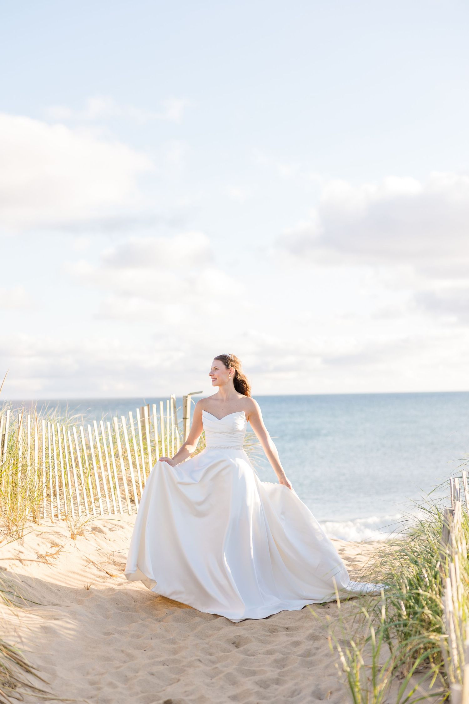 Elegant bride in a flowing strapless white gown stands on a sandy beach path with the ocean behind her.