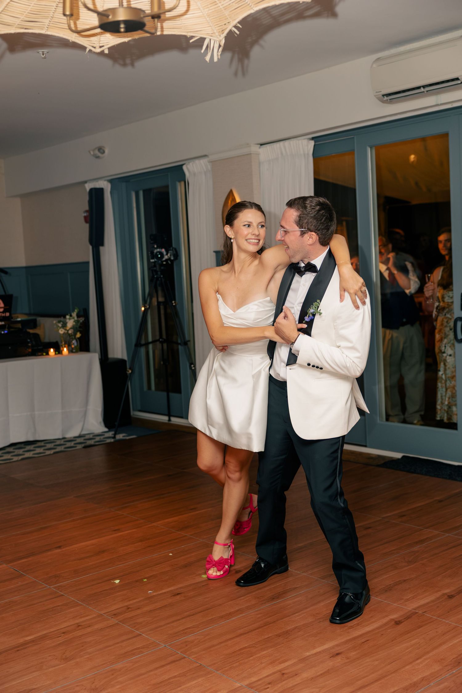 Newlyweds share first dance in white tuxedo and short white dress with pink heels at wedding reception.