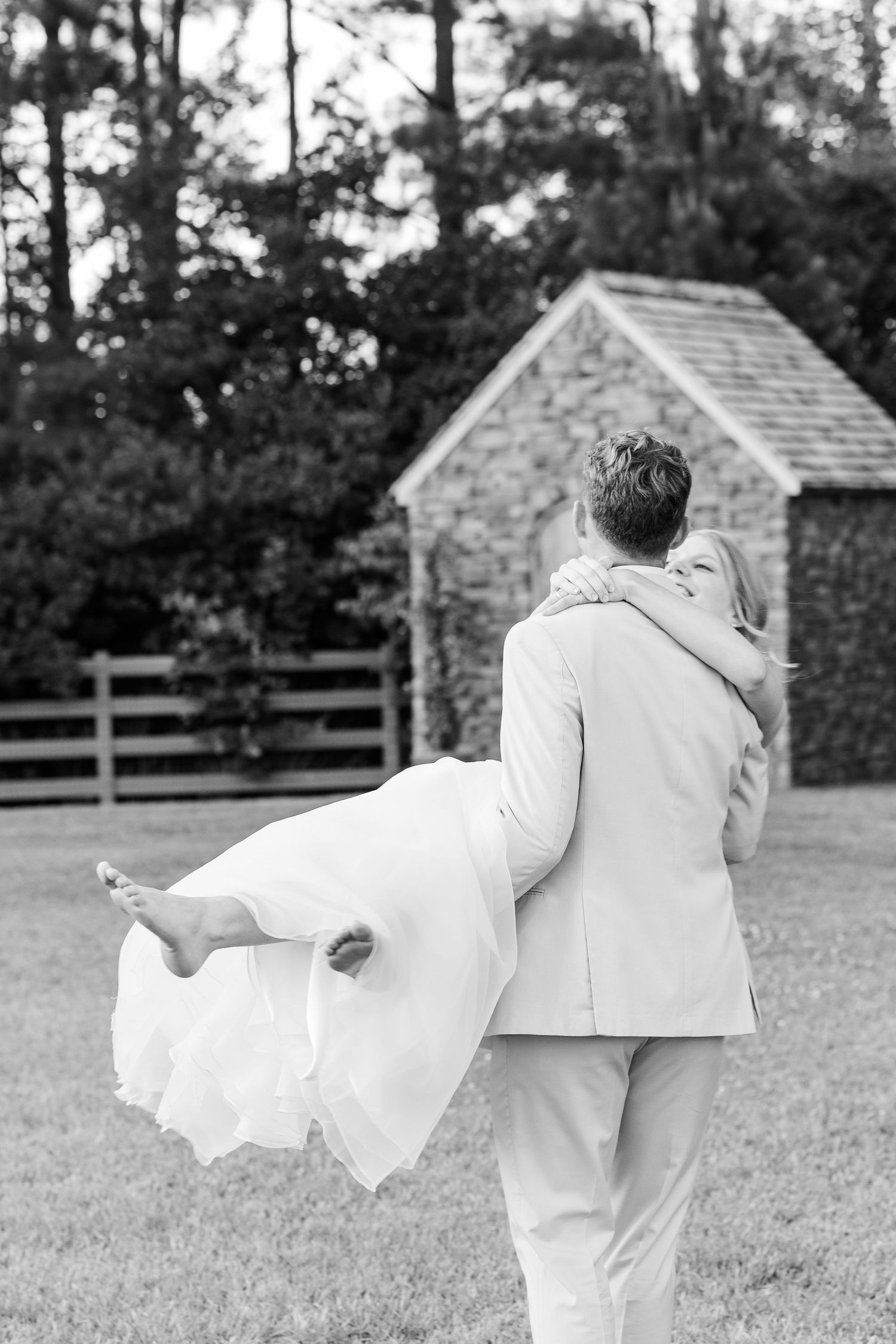 A couple in all white attire playfully dances together in a grassy field, creating a romantic black and white scene.