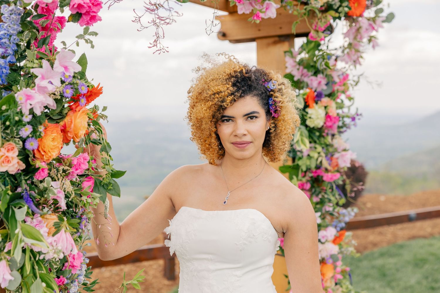 Close-up of bride with curly hair and flower crown standing beside vibrant colorful floral wedding arch.
