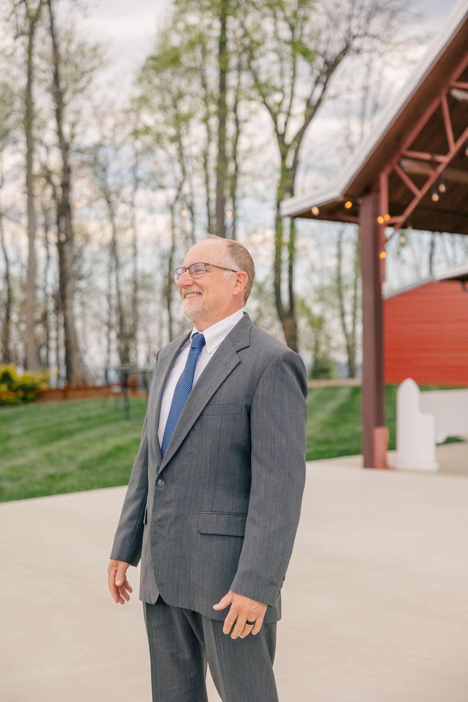 Smiling older man in gray pinstripe suit with blue tie standing outdoors at wedding venue.