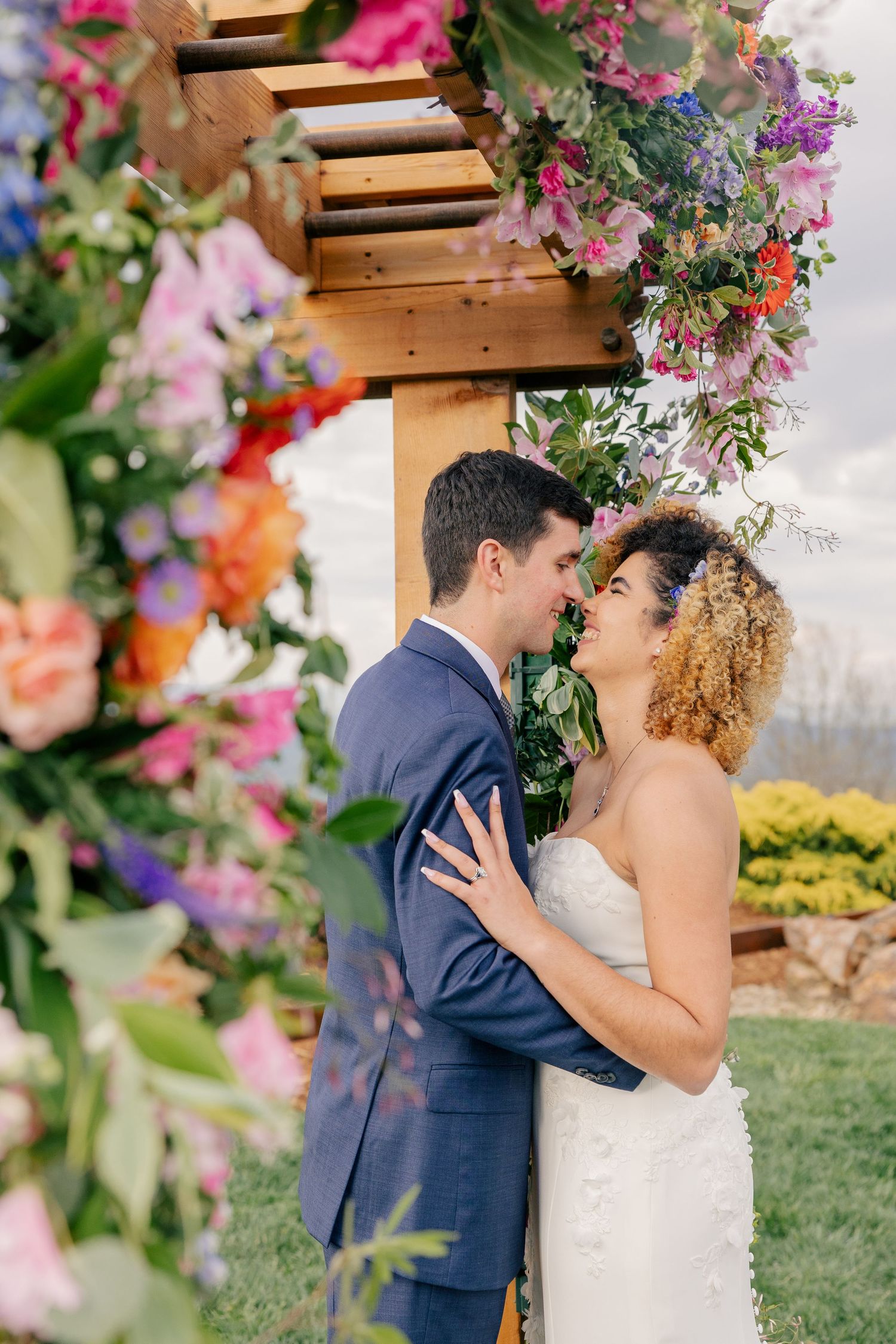 Bride and groom sharing a romantic moment under a vibrant floral wooden arch at their outdoor ceremony.