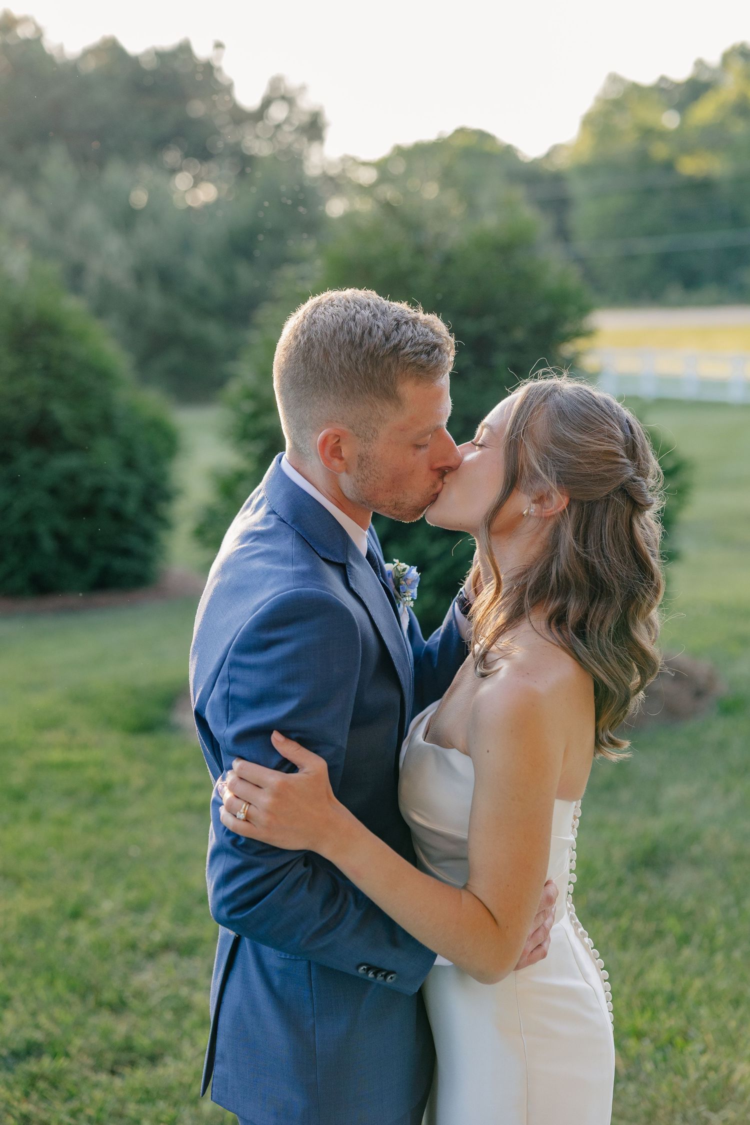 Bride and groom share romantic golden hour kiss outdoors surrounded by lush green garden landscape.