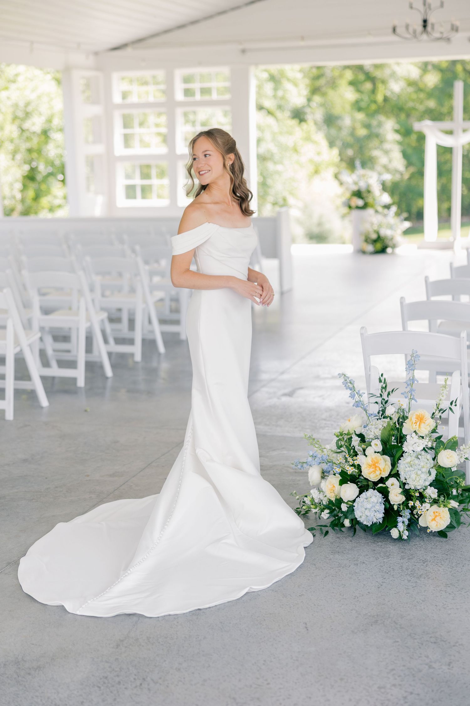 An elegant bride in a white off-shoulder fitted gown with a train stands in a sunlit outdoor ceremony venue.