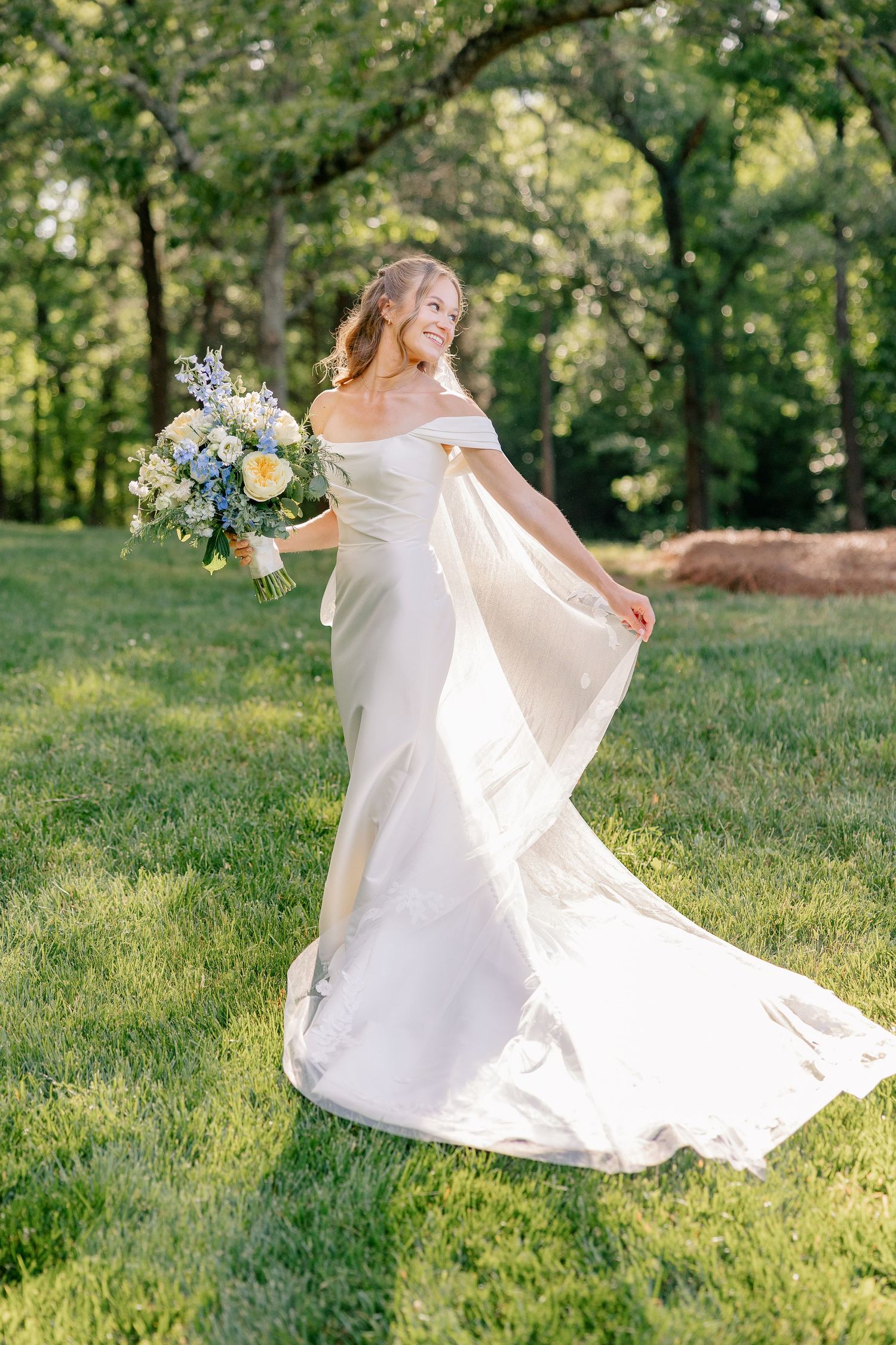 Radiant bride in off-shoulder white gown twirls with blue and white bouquet on sunlit green lawn.