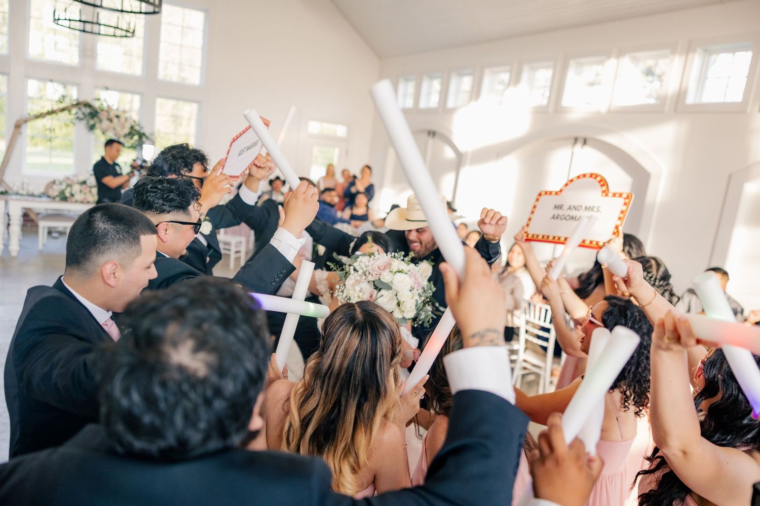 Wedding reception guests celebrating with hands raised in joyful dance at bright venue.