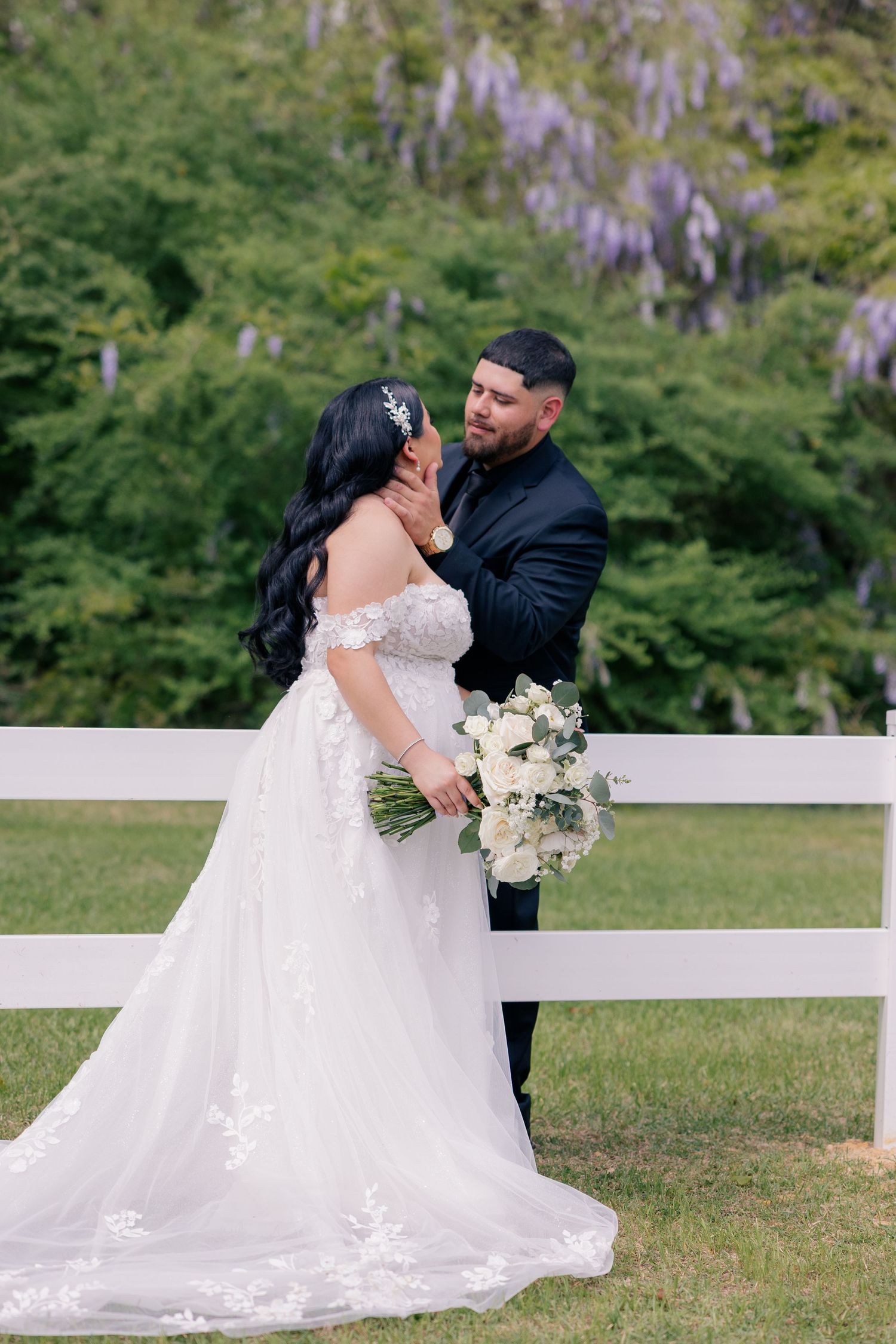 Couple in elegant wedding attire share romantic moments on a lawn with blooming wisteria.