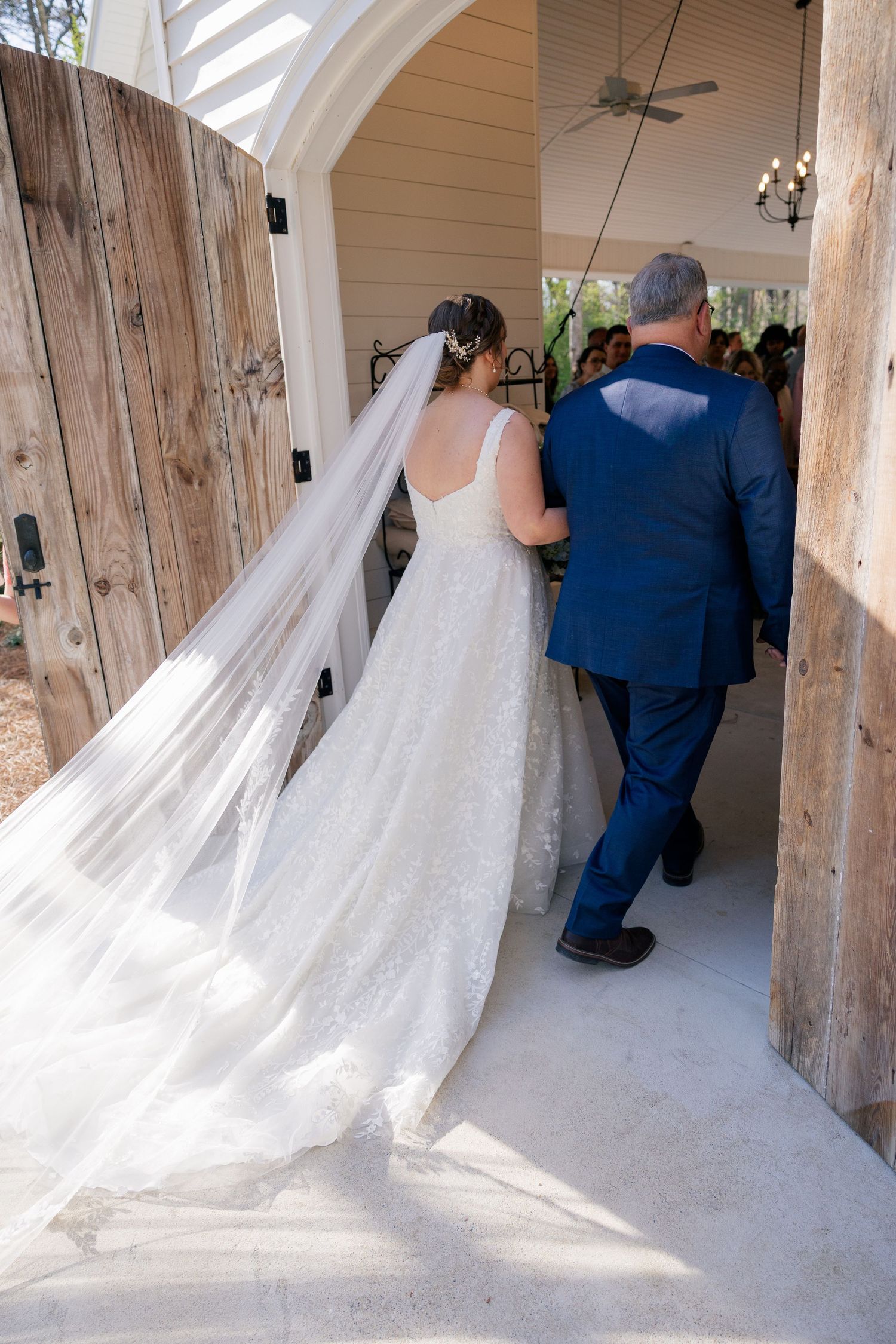 A bride in a flowing white gown with long veil enters a rustic chapel doorway.