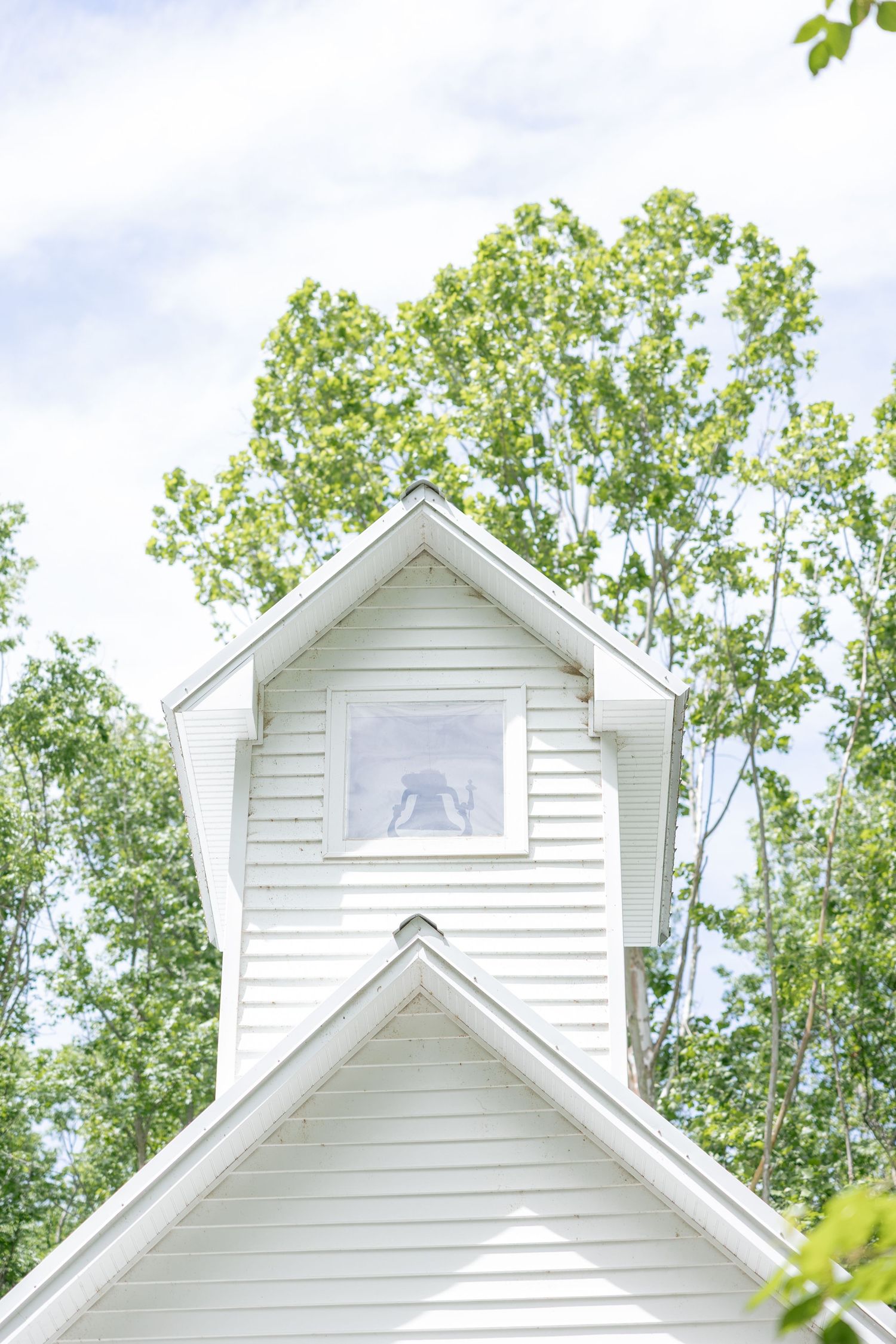 White chapel steeple with small windows and gable roof against a backdrop of green trees and blue sky.