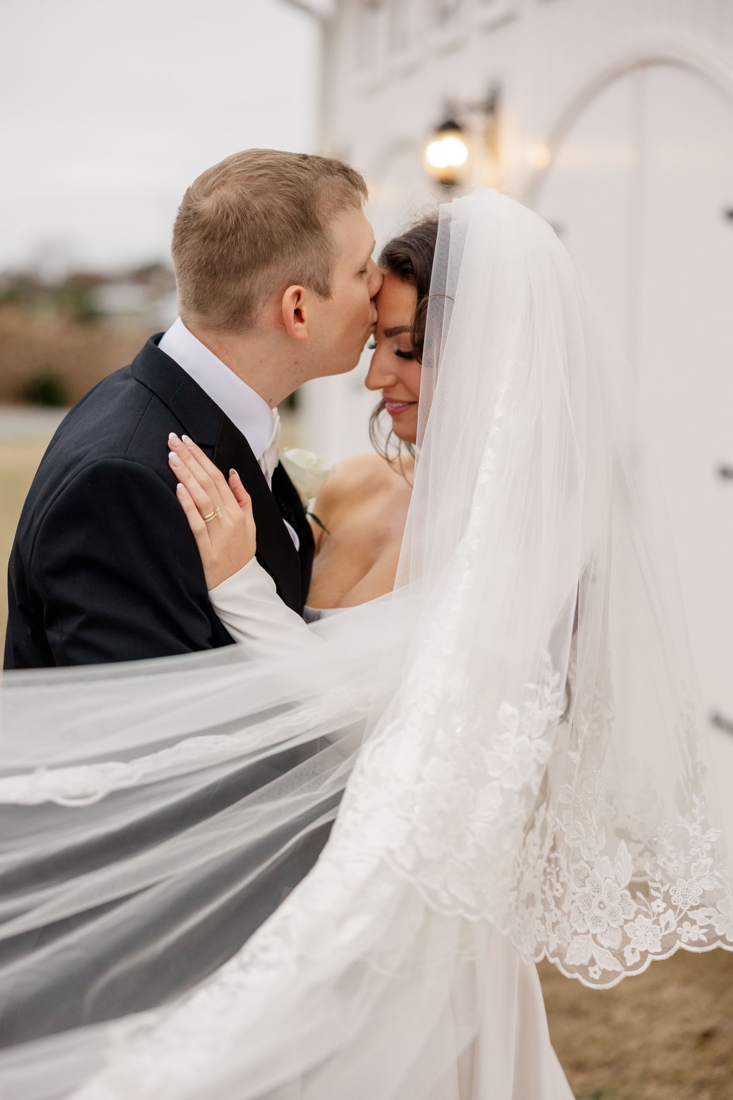 Romantic wedding couple embraces under flowing white veil against church exterior.
