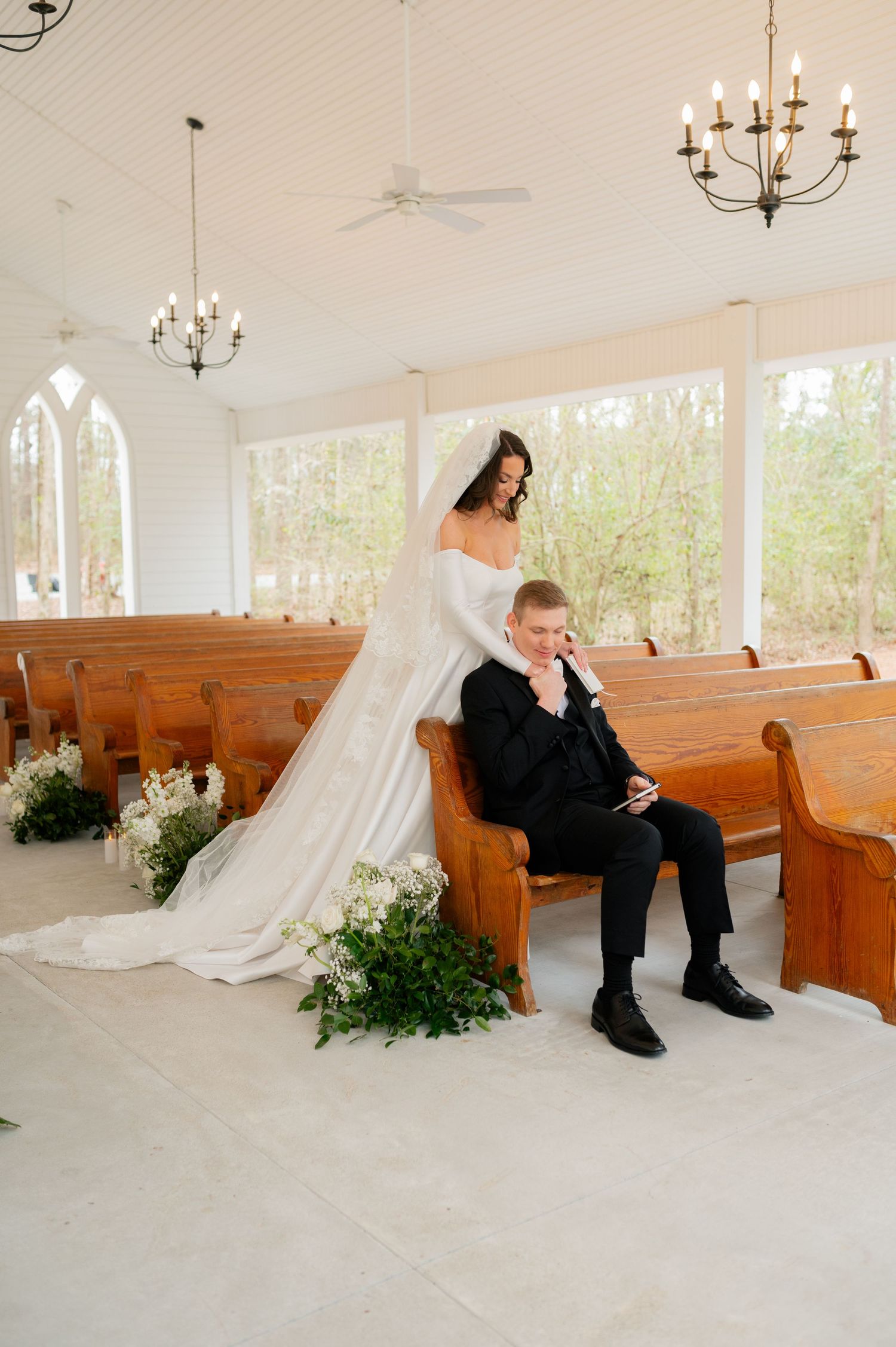 Bride and groom sitting separately on wooden church pews in an elegant white chapel.
