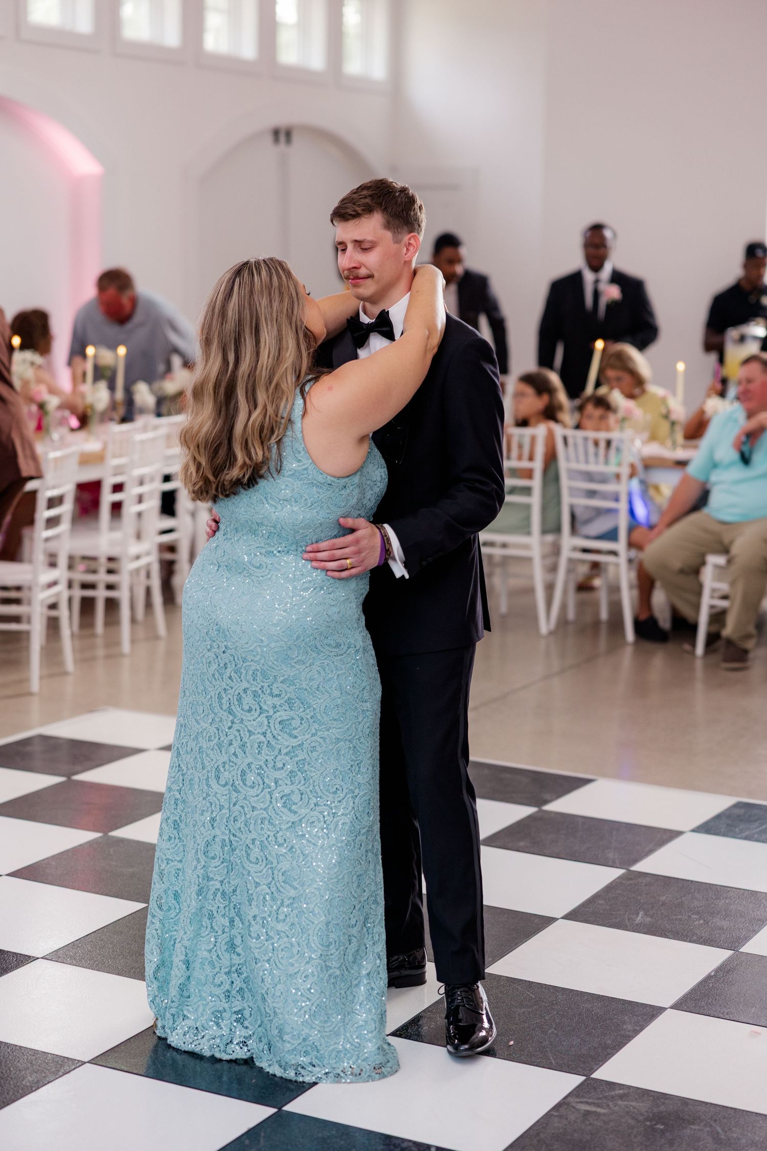 A couple dances together on a black and white checkered floor at a wedding reception with white chairs in the background.