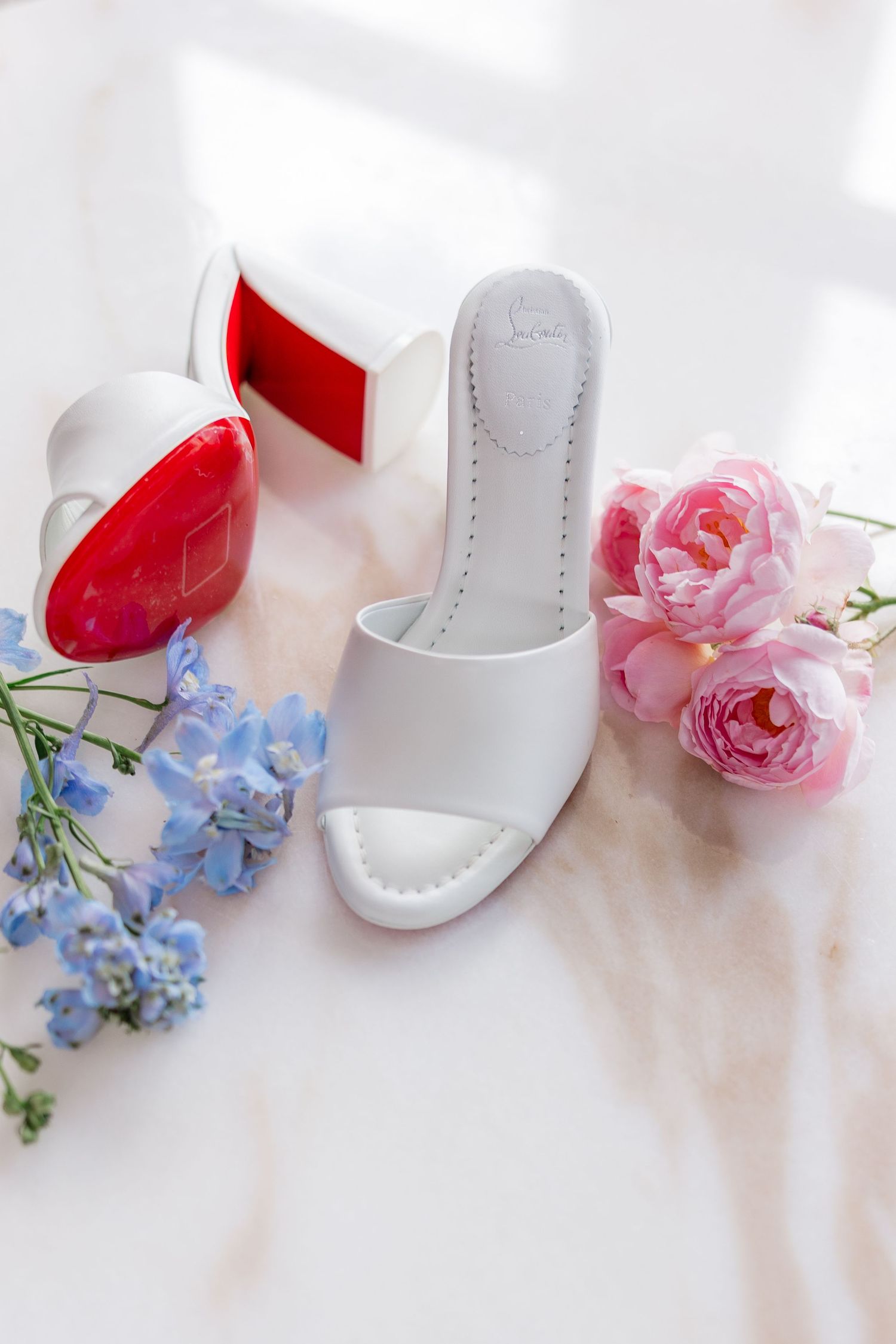 White wedding shoes arranged with delicate pink roses and blue hydrangeas against a light marble background.