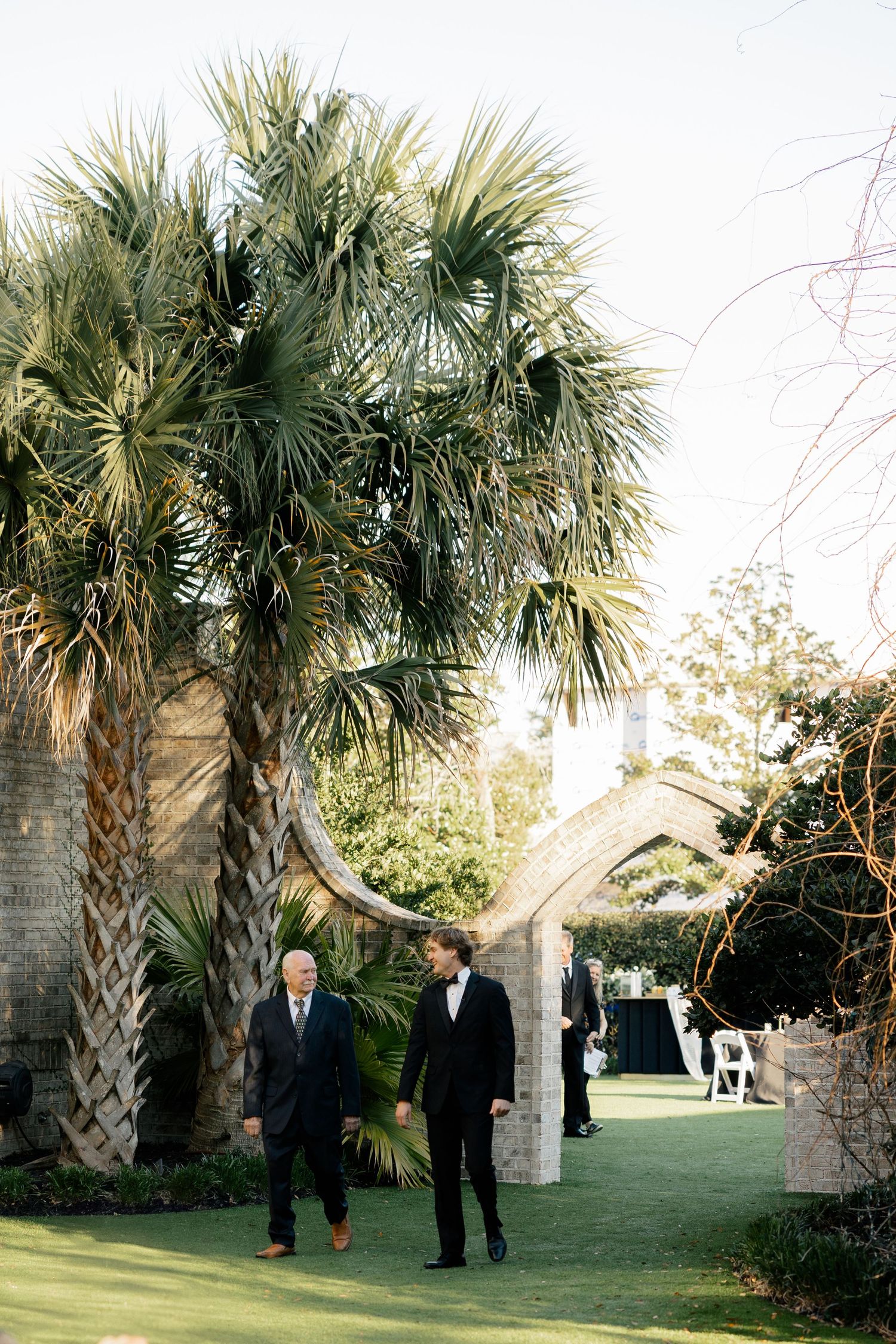 People walking through a garden path lined with tall palm trees and stone archways.