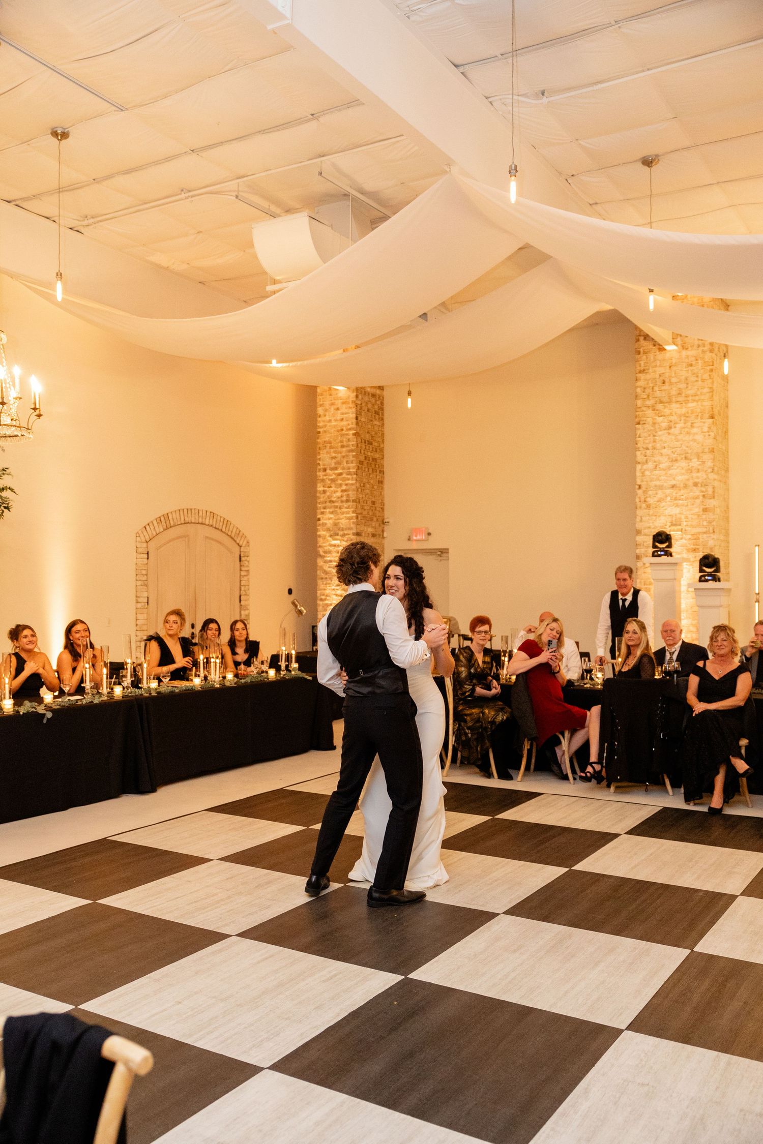 A couple shares their first dance on a black and white checkered floor under draped fabric and chandeliers.