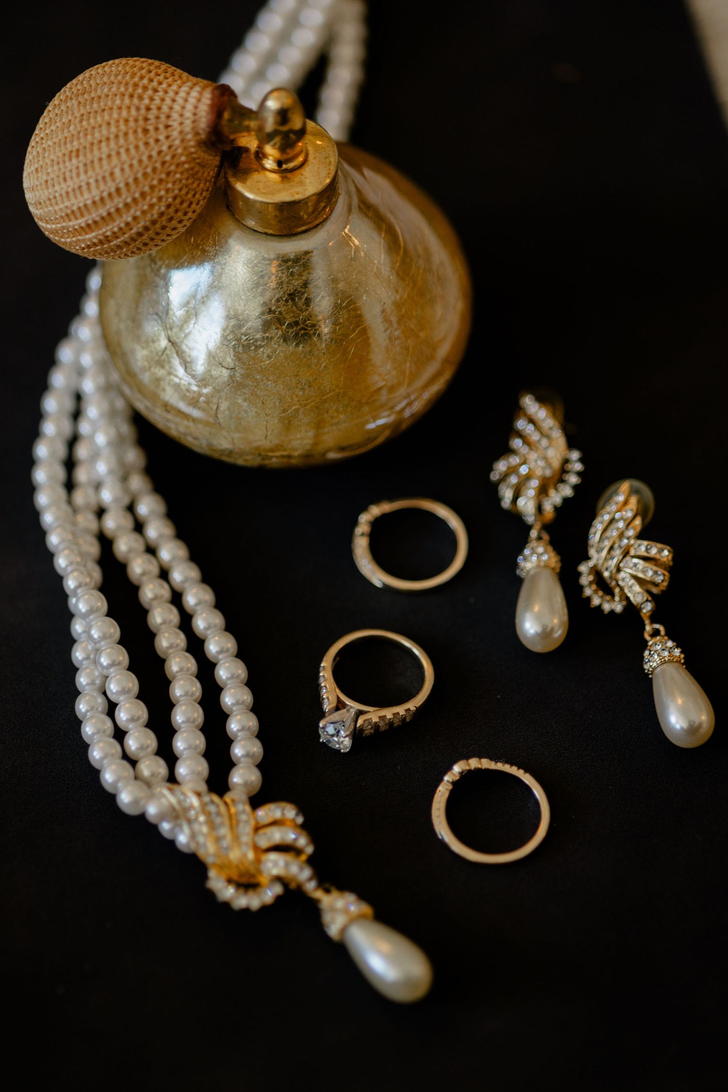 Elegant bridal accessories including pearl necklace, rings, earrings and vintage perfume atomizer on black background.