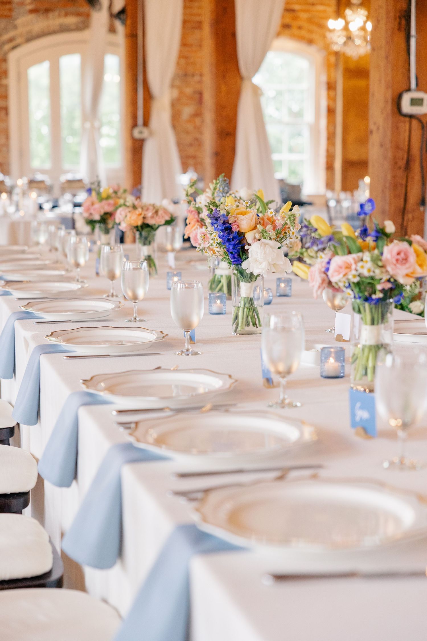 Elegant wedding reception table set with blue napkins and colorful floral centerpieces in brick venue.