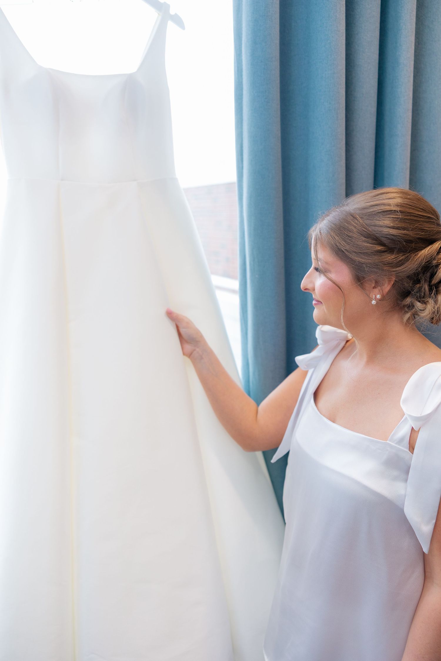 Bride admires her white wedding gown hanging by blue curtained window before ceremony.