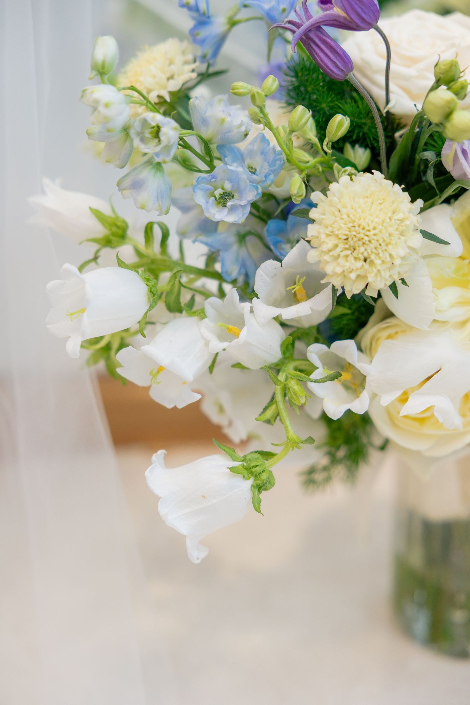 Elegant wedding bouquet featuring white snapdragons, blue flowers, cream scabiosa, and lush greenery in a glass vase.