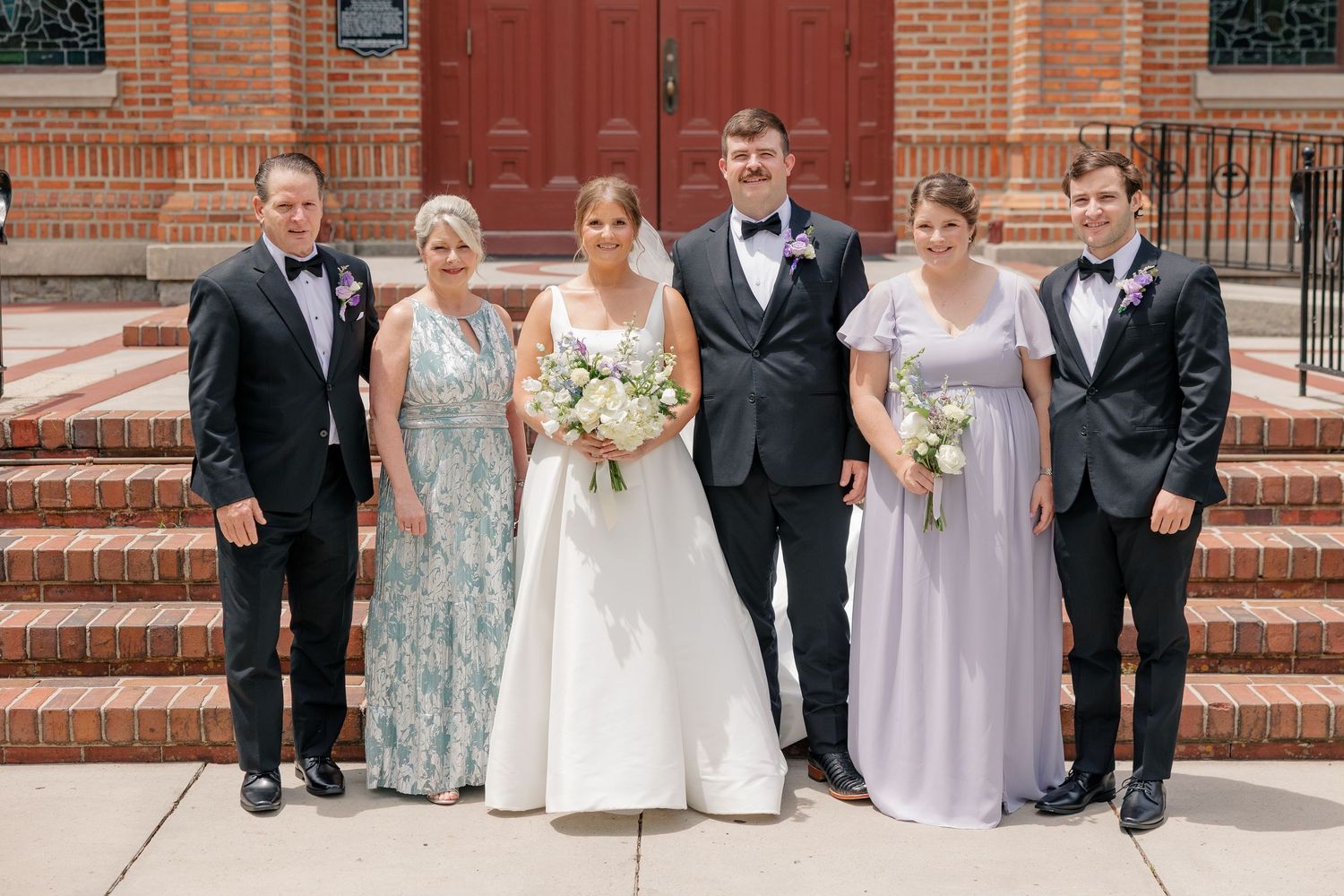 Wedding party of six poses on church steps with bride in white gown and bridesmaids in lavender dresses.