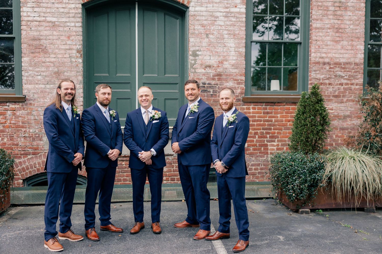 Five groomsmen in matching navy blue suits with boutonnieres pose smiling outside a brick building with green doors.