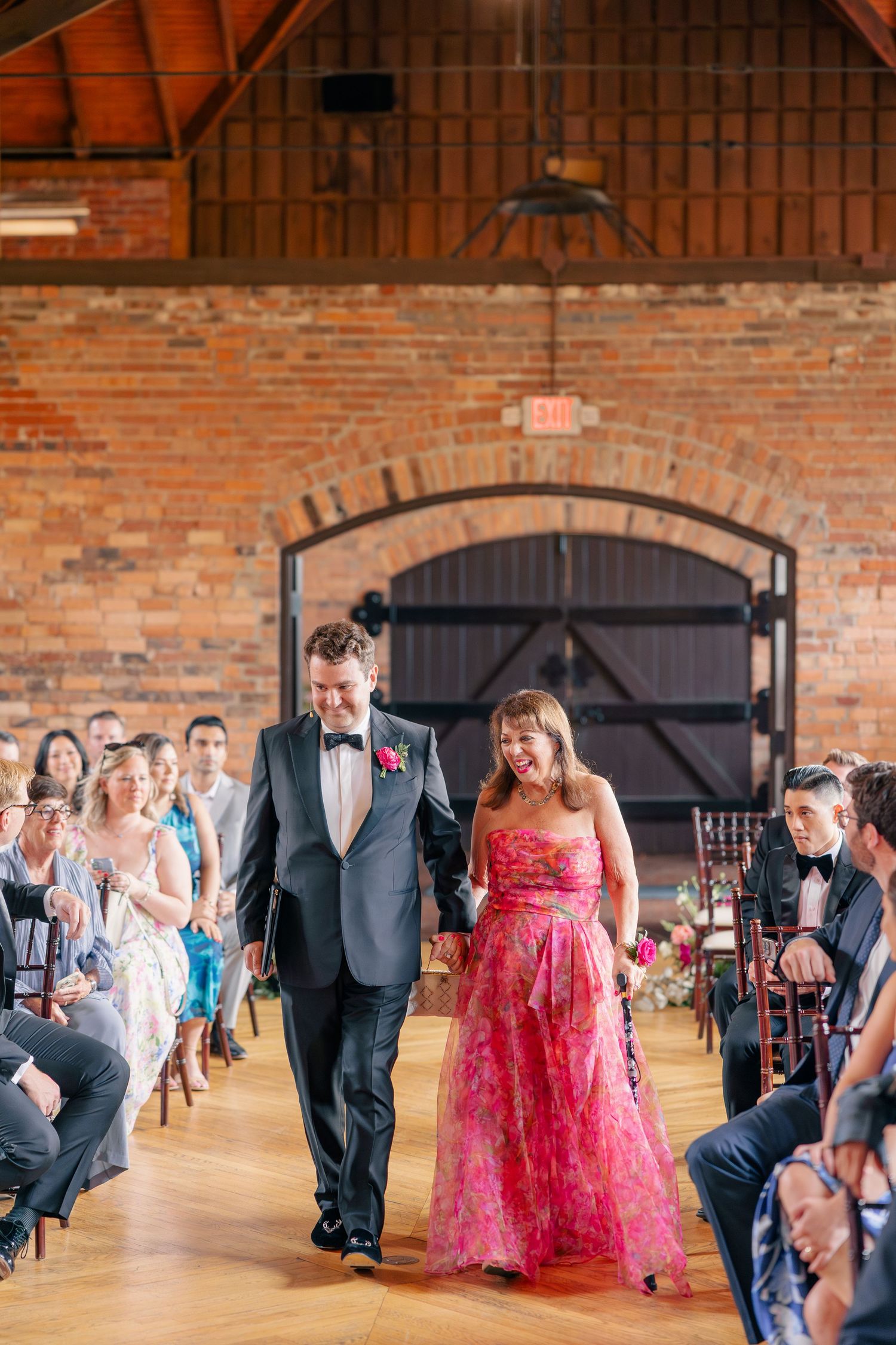 Groom escorts smiling woman in pink floral gown down aisle in rustic brick ceremony venue.