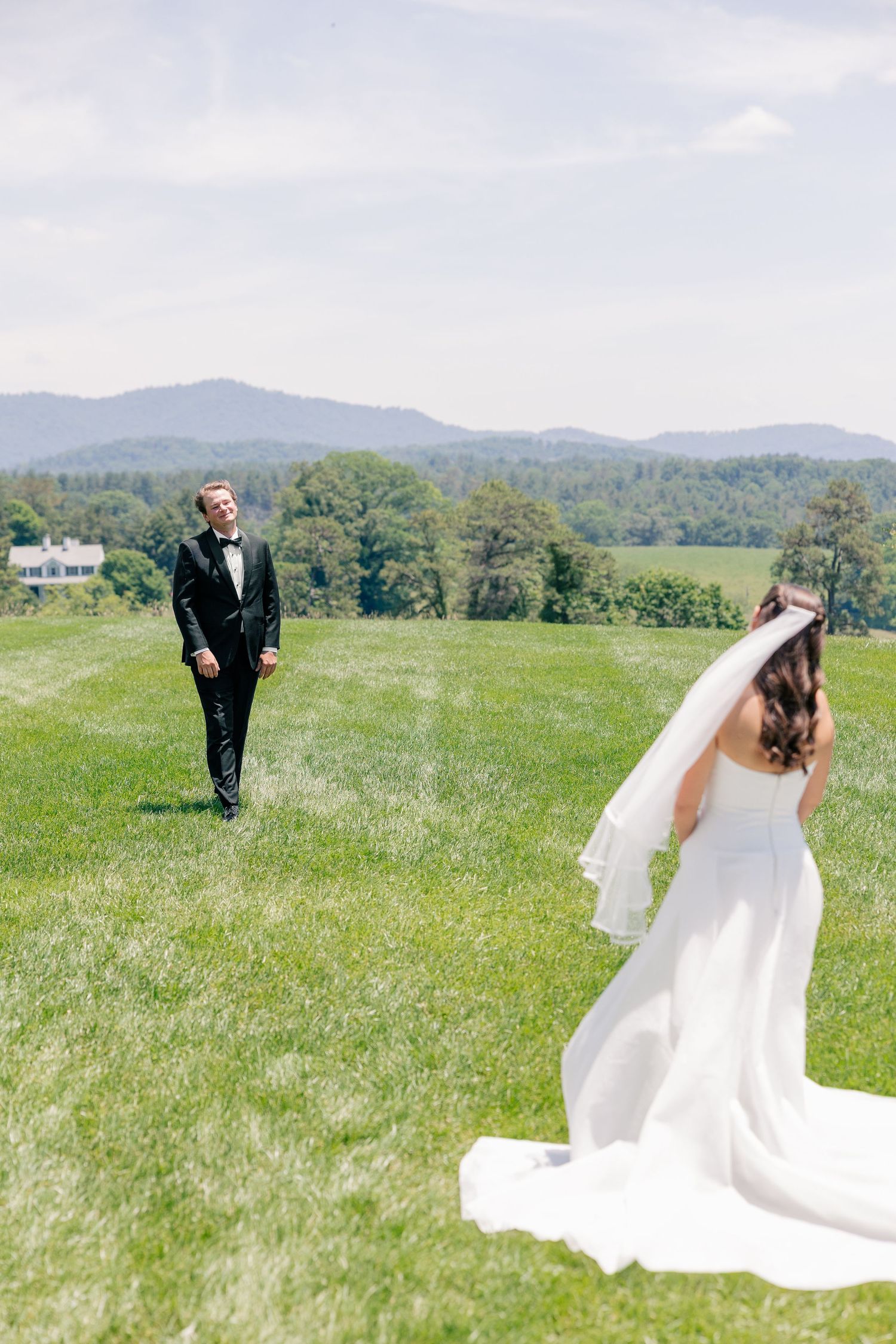 Bride in flowing white gown and groom in tuxedo sharing first look on green mountain meadow.