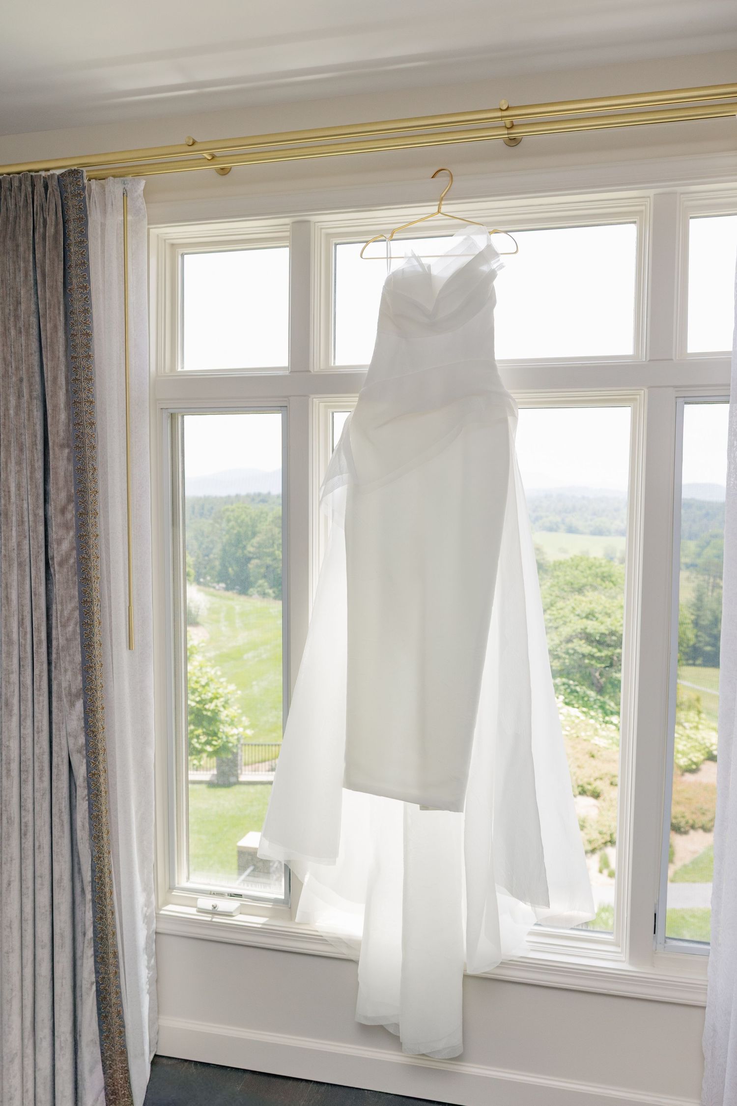 Elegant white wedding dress hanging on hanger in sunlit window with scenic view.