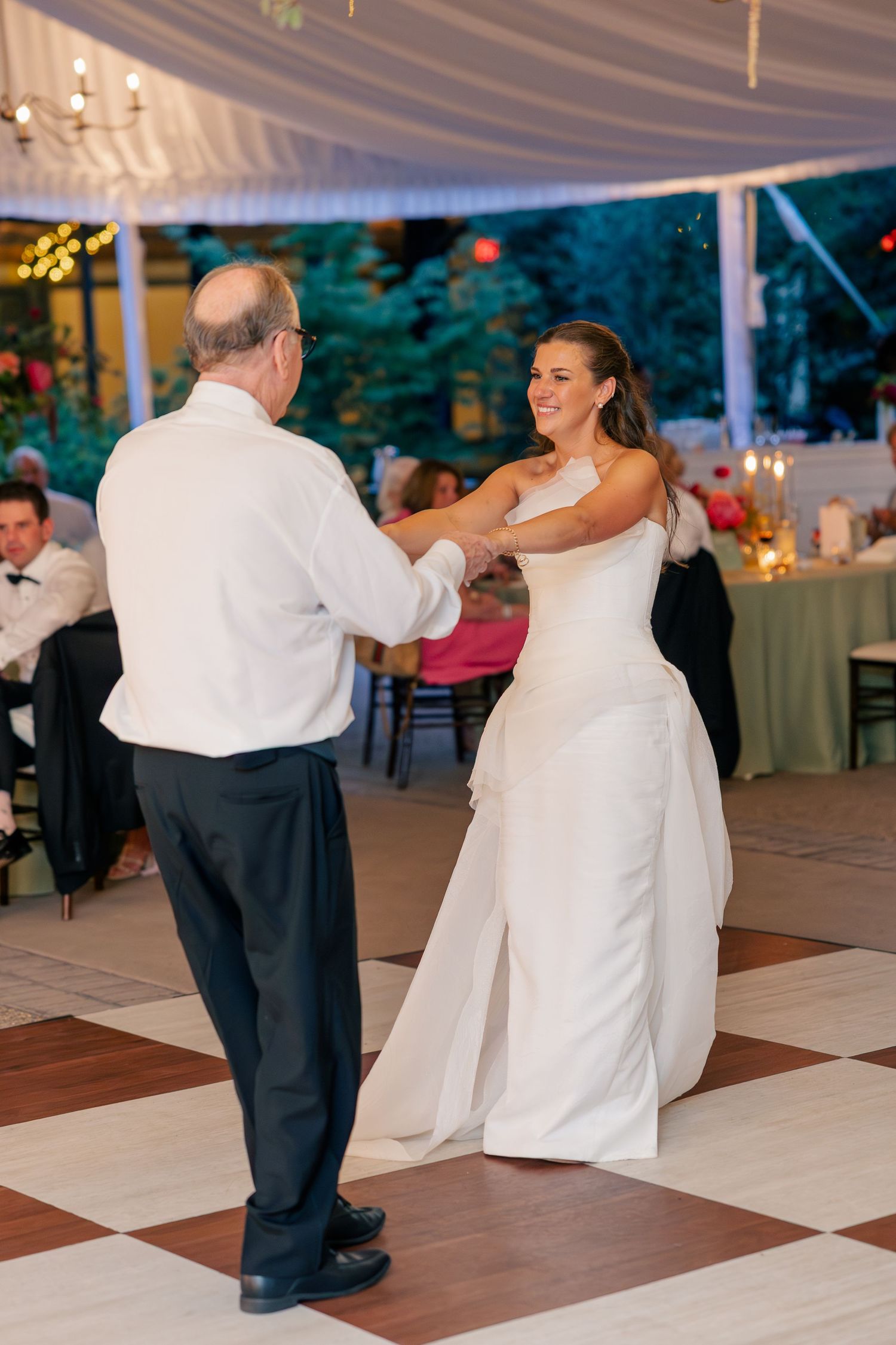 Bride in white gown shares father-daughter dance under elegant draped tent at wedding reception.